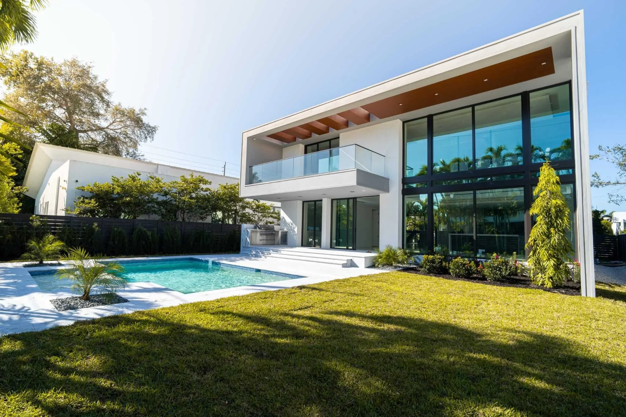 Modern white house with large glass windows, a balcony, and a pool in the backyard, surrounded by green grass and trees under a clear blue sky.