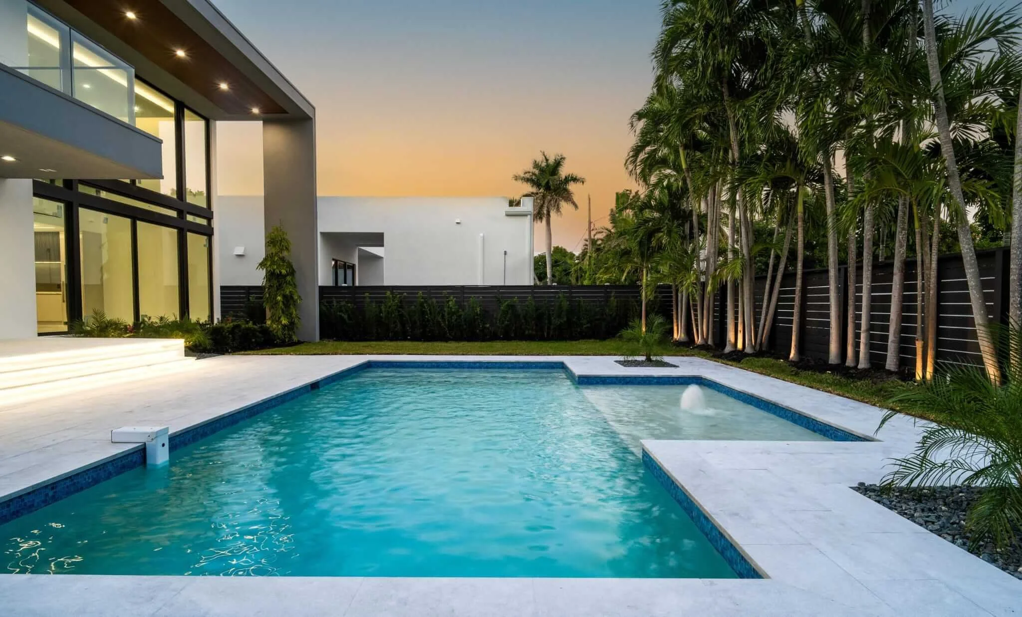 Modern backyard with a swimming pool, surrounded by tropical palm trees, a patio area, and a contemporary house with large glass windows during sunset.