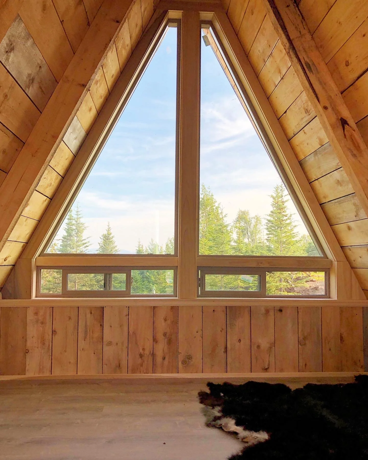 A triangular-shaped window in a wooden room, showing a view of trees and a blue sky with clouds outside.