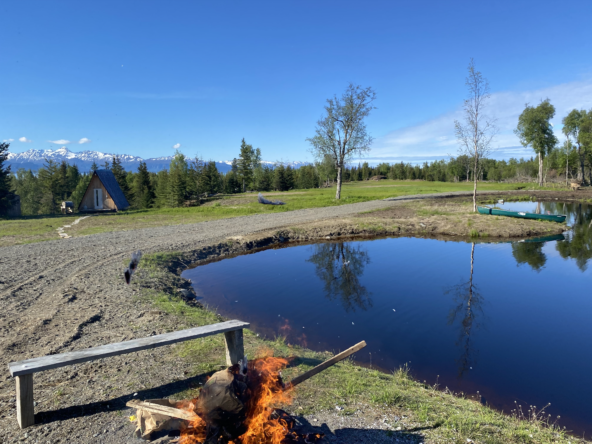 Scenic view of a rural landscape with a small cabin, a pond, trees, and snow-capped mountains in the background under a blue sky, with a campfire in the foreground.