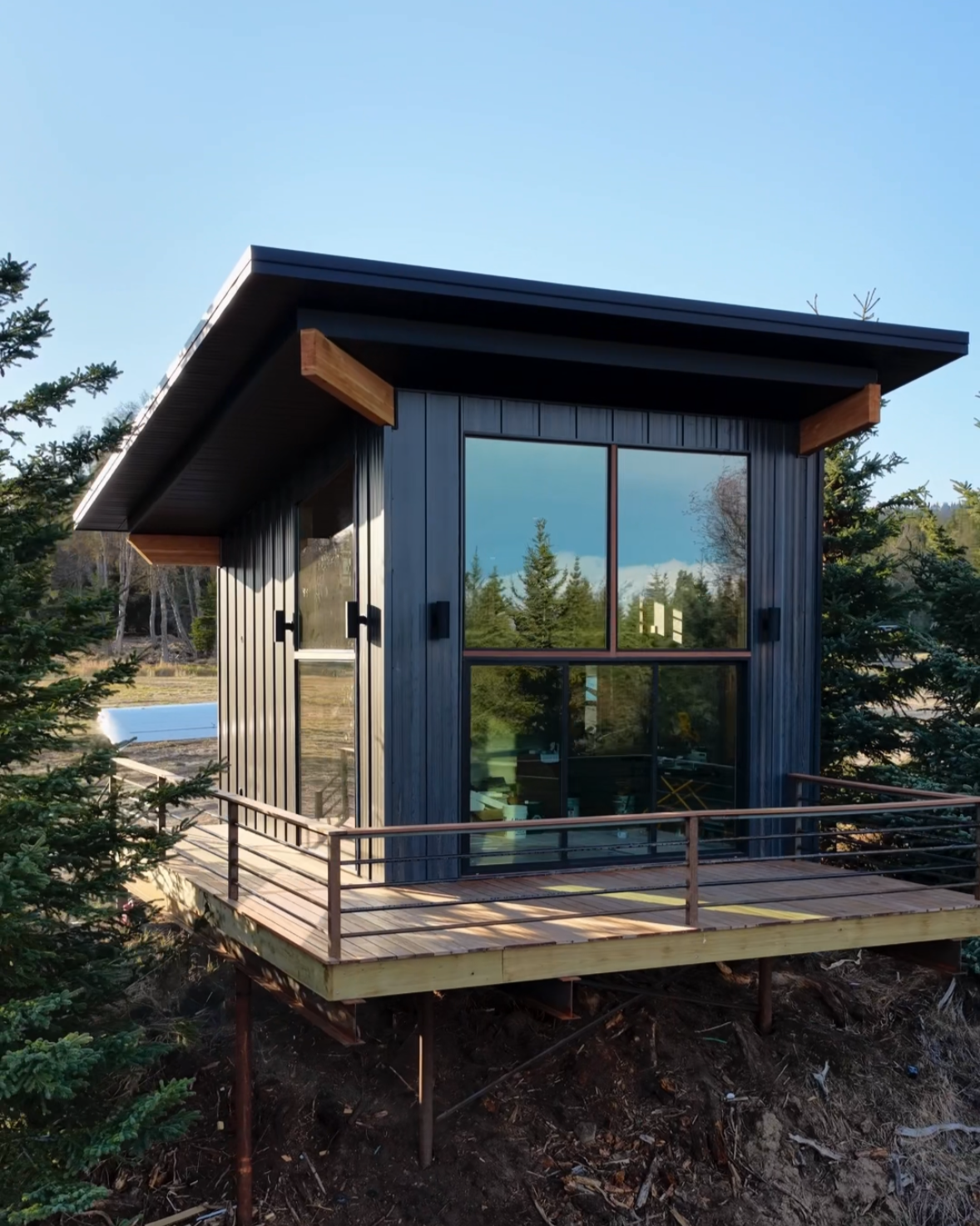 A small wooden A-frame cabin with a black metal roof, a white door with a vertical window, and a small wooden porch with chains on either side, surrounded by trees and blue sky.