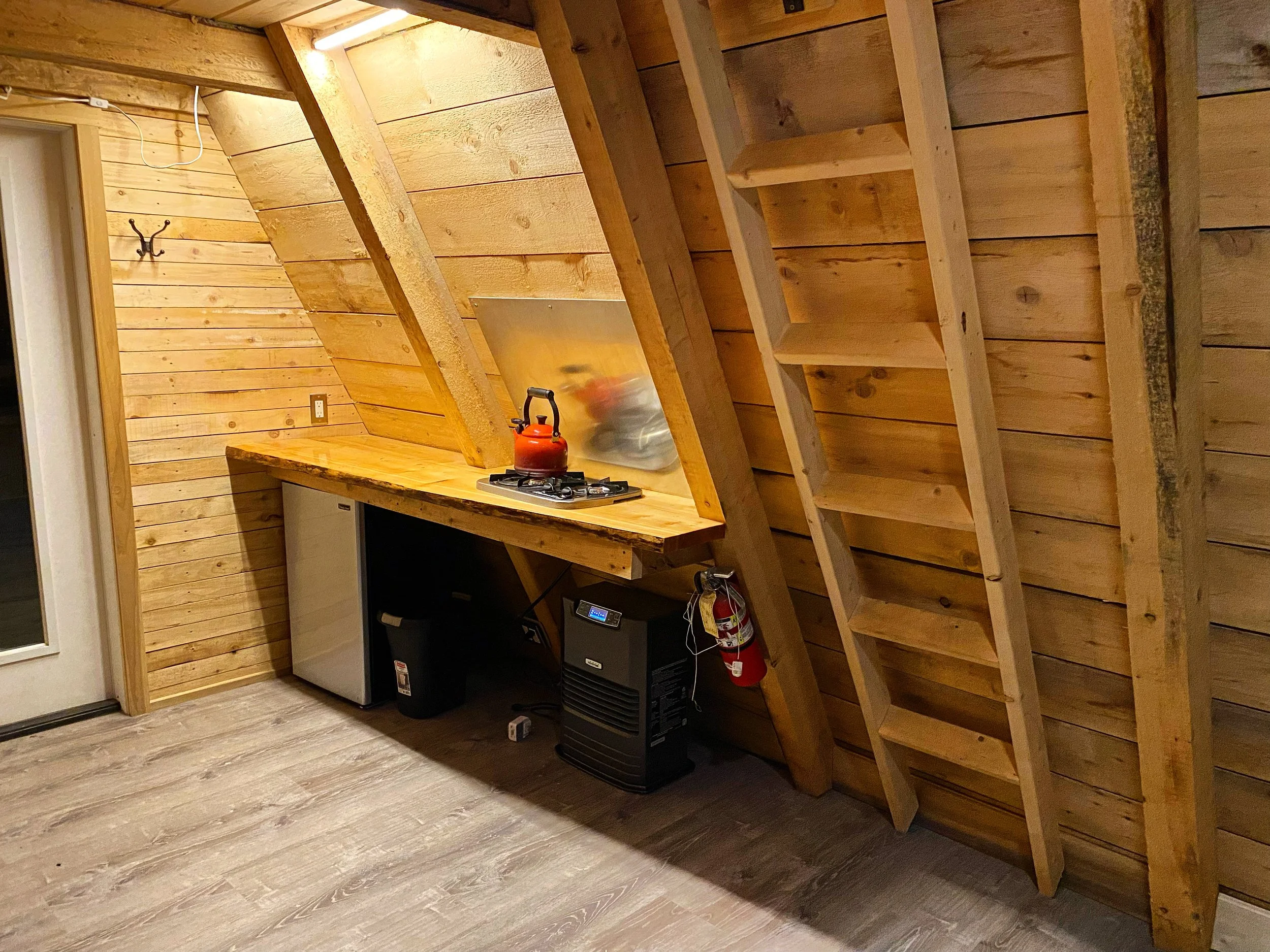 A small kitchen area with wood-paneled walls and sloped ceiling. There is a wooden countertop with a red kettle on a gas stove, a mini fridge, and a portable heater below the counter. A ladder is leaning against the wall, and a fire extinguisher is m