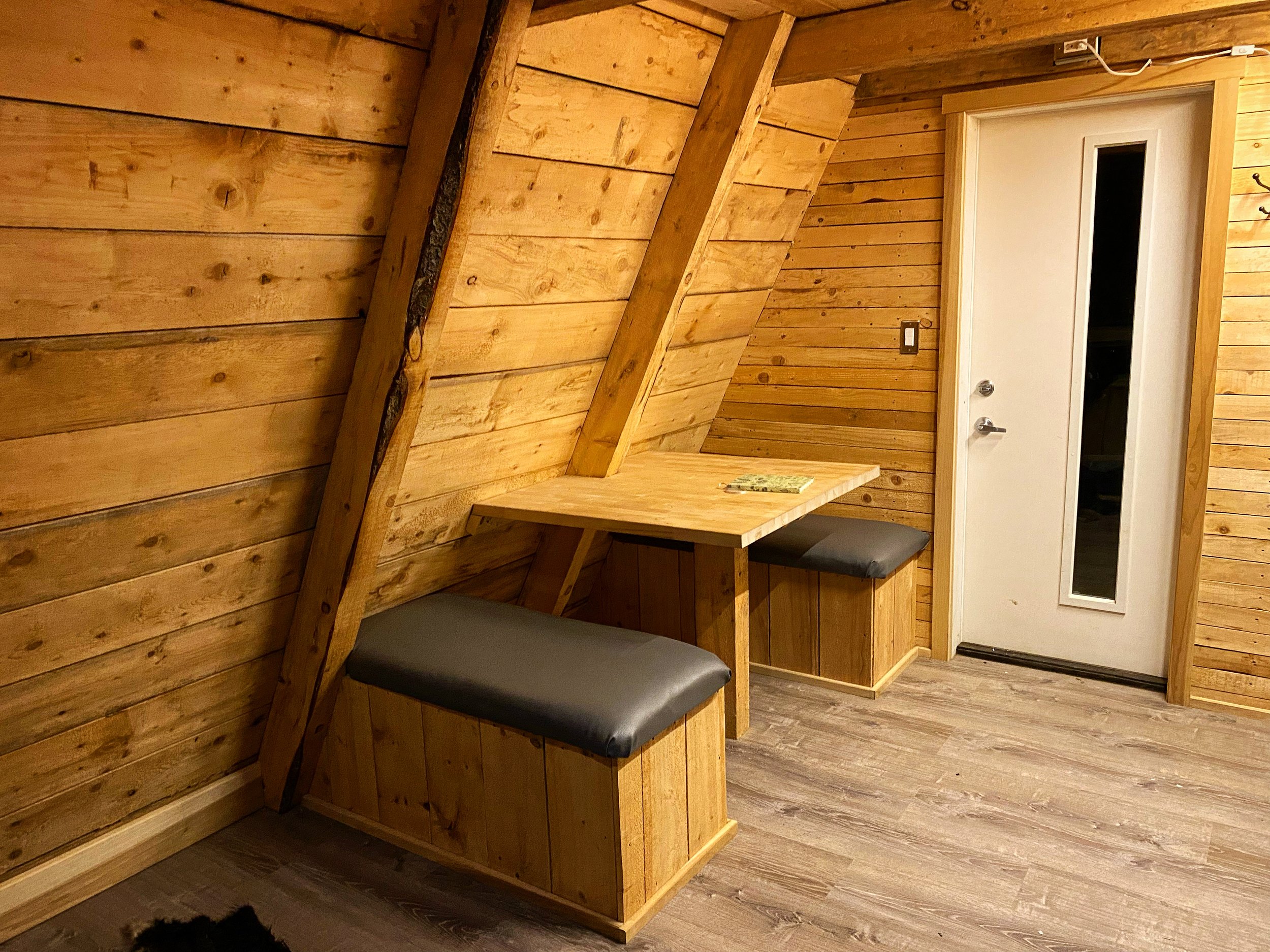 A corner of a wooden interior with slanted walls, a small wooden table with seating, and a white door with a window.