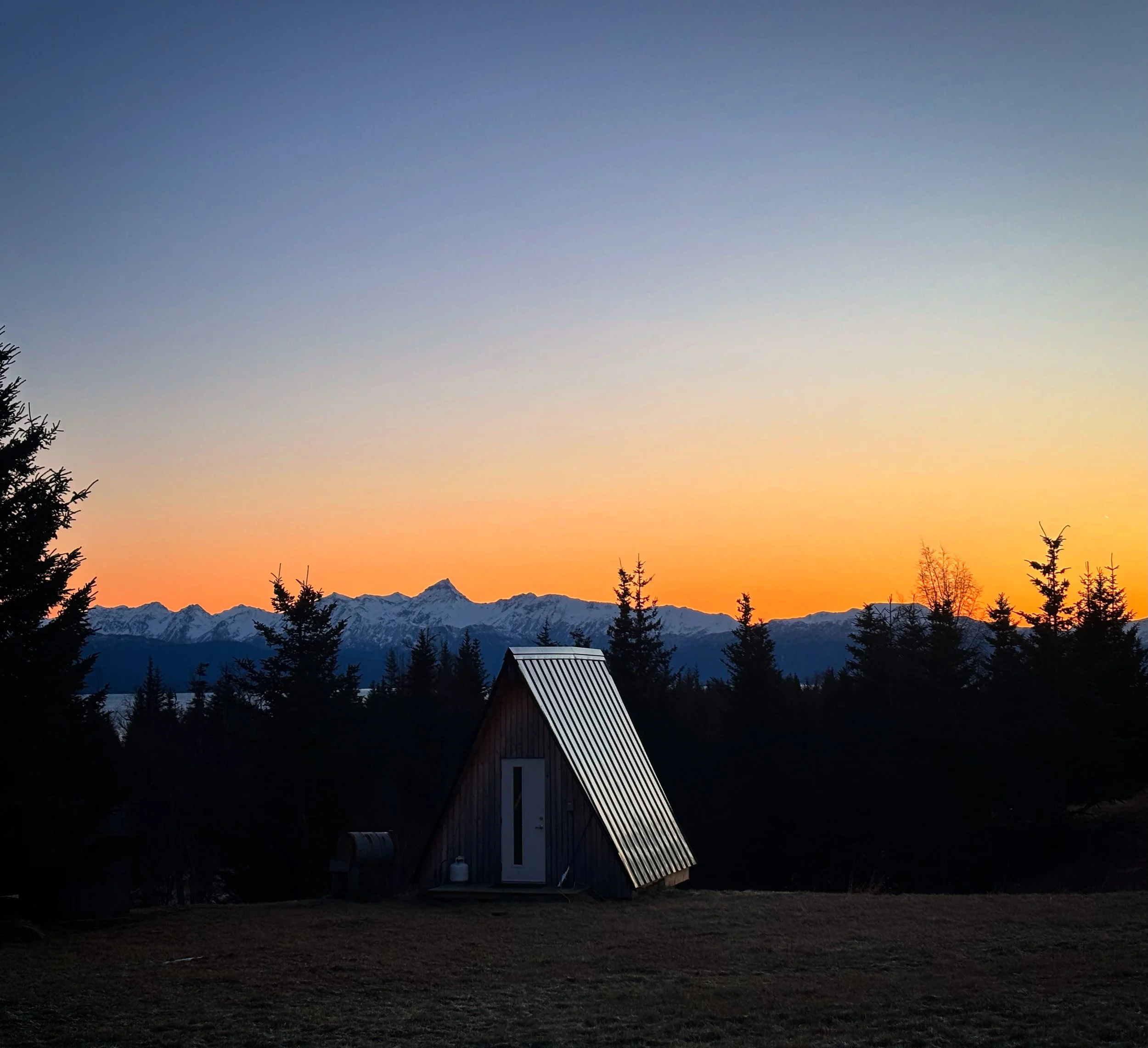 A small A-frame cabin with metal roof, set on a grassy area surrounded by trees, with snow-capped mountains in the background under a sunset sky.