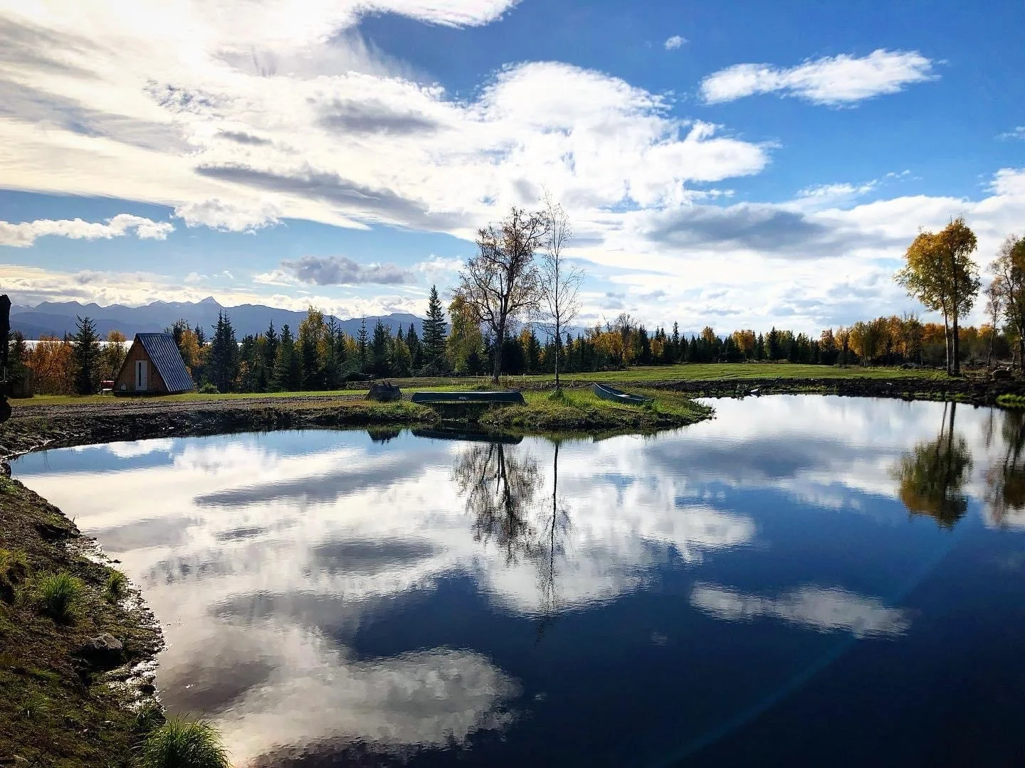 A peaceful rural scene with a calm river reflecting the partly cloudy sky, trees with autumn foliage, and small boats near the riverbank.