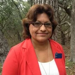 A woman with short brown hair and glasses wearing a red blazer and a white top, standing outdoors with trees in the background.