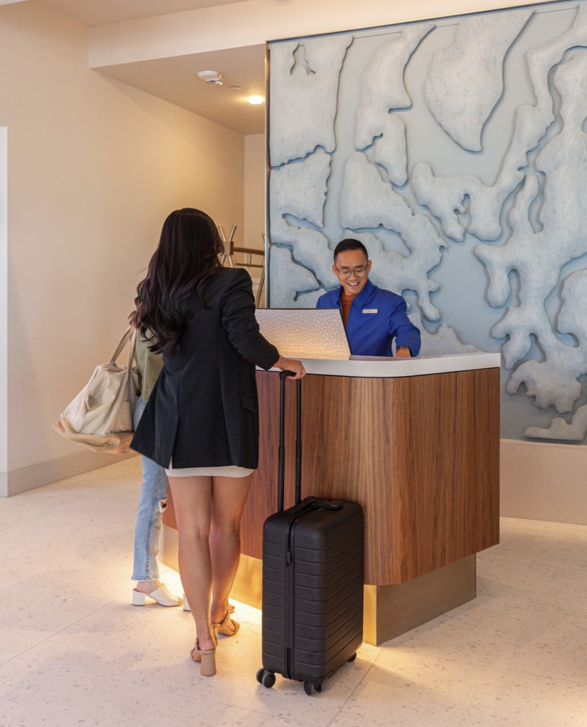 Hotel front desk receptionist smiling while assisting a guest with a rolling suitcase.