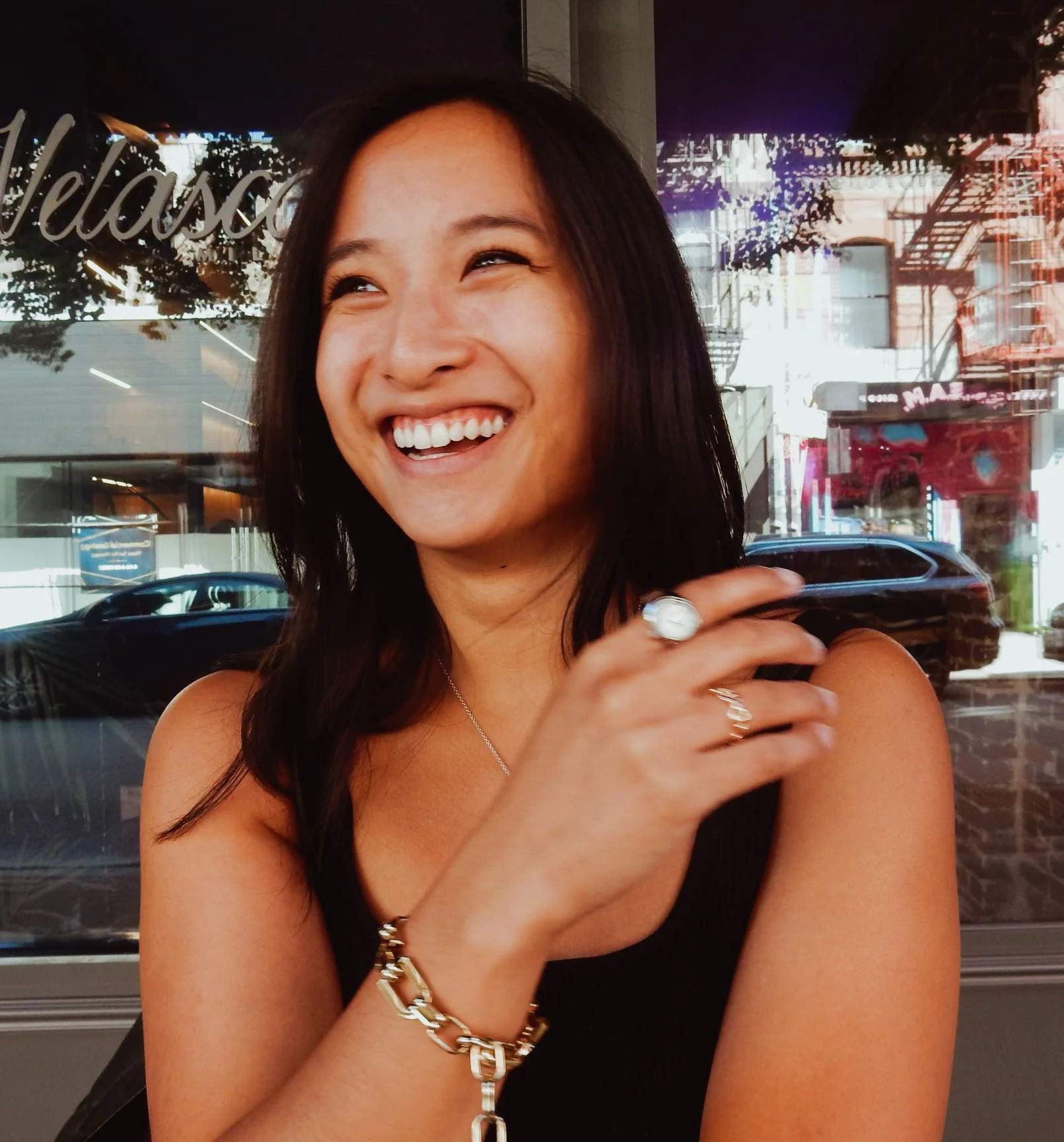A woman with long dark hair smiling and looking to the side inside a shop or cafe.