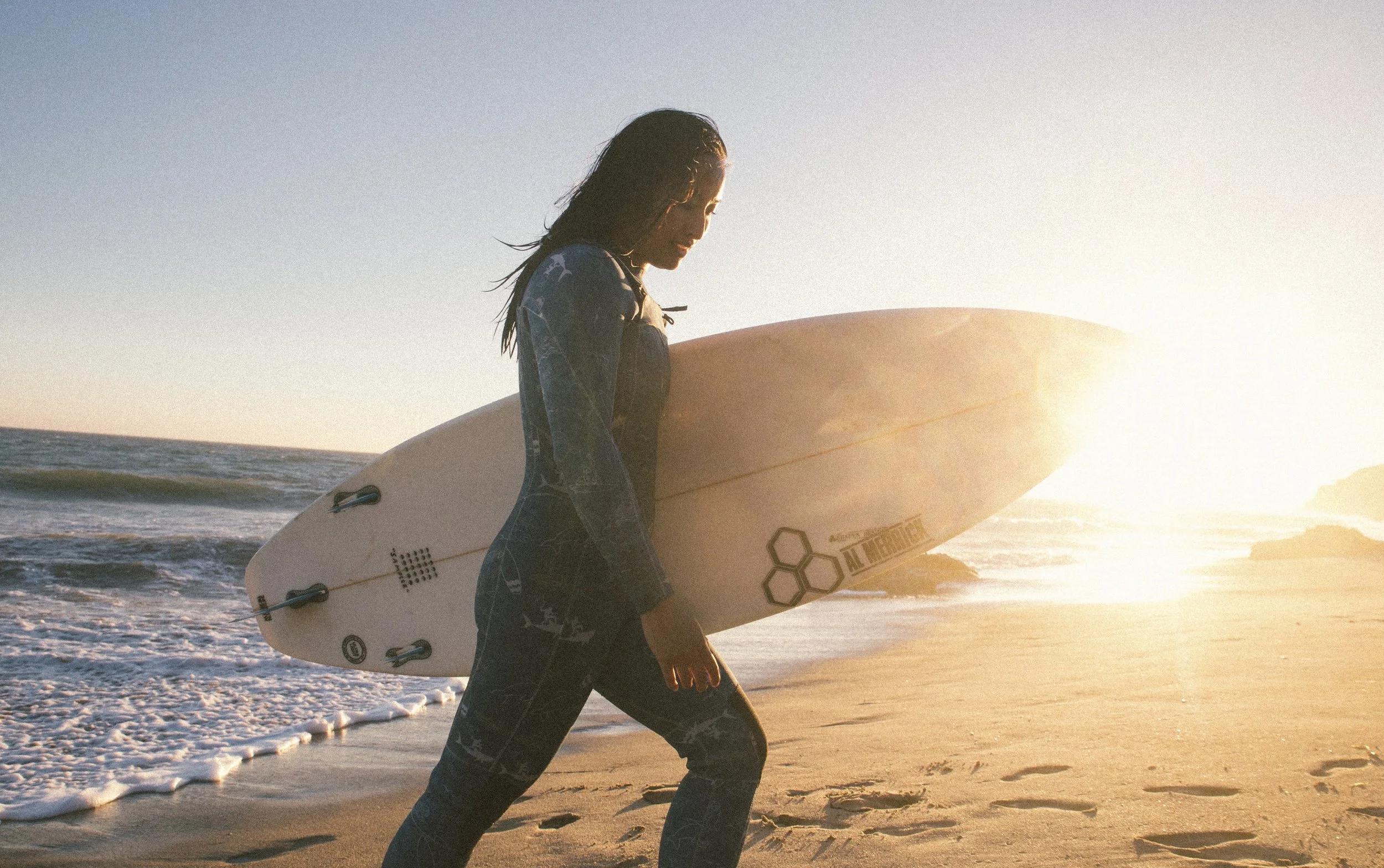 A woman walking on the beach holding a surfboard at sunset.