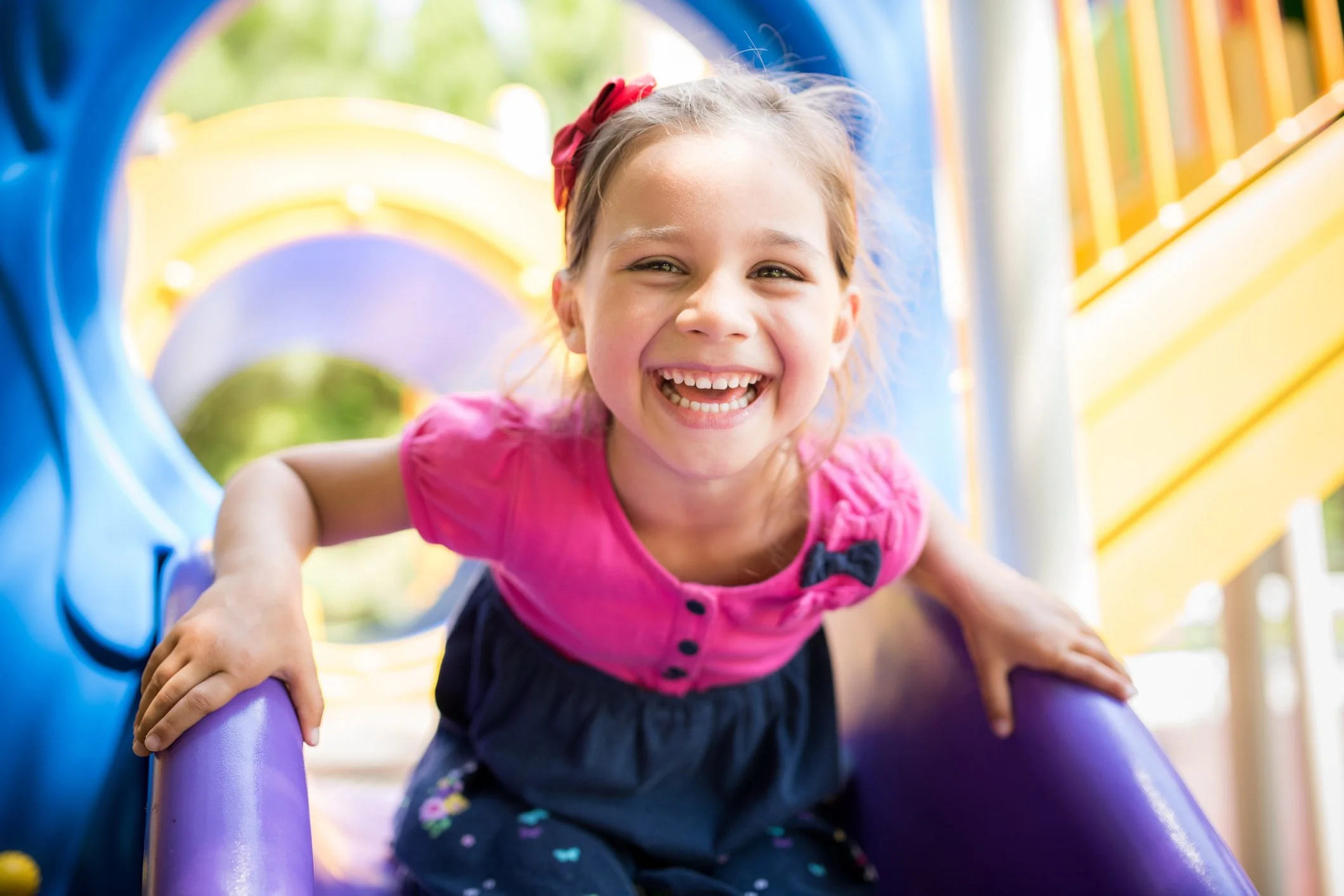 A young girl with a big smile, wearing a pink top and navy skirt, playing on a colorful playground slide.