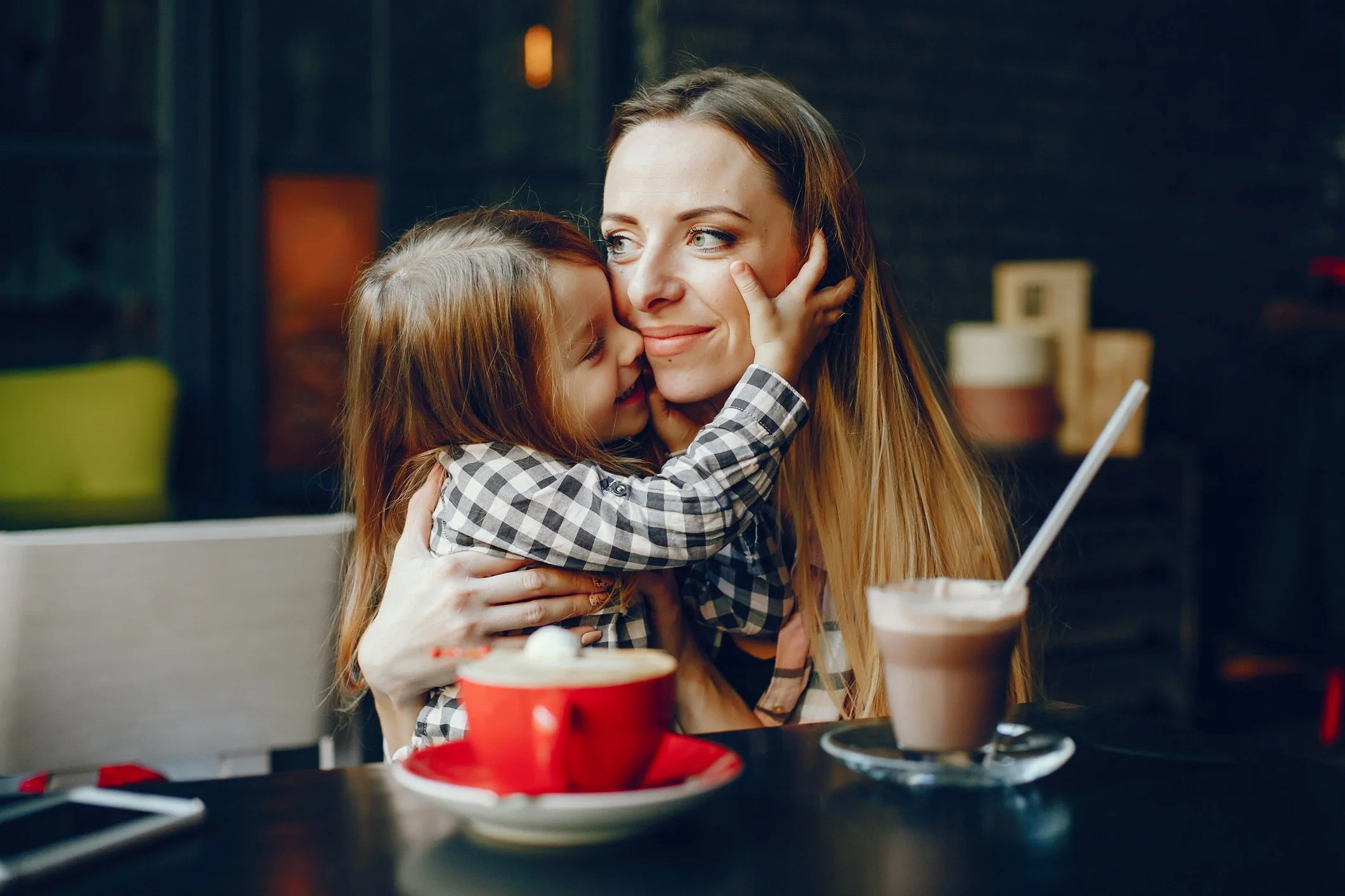 A woman and a young girl sharing an affectionate hug at a cafe, with drinks on the table.
