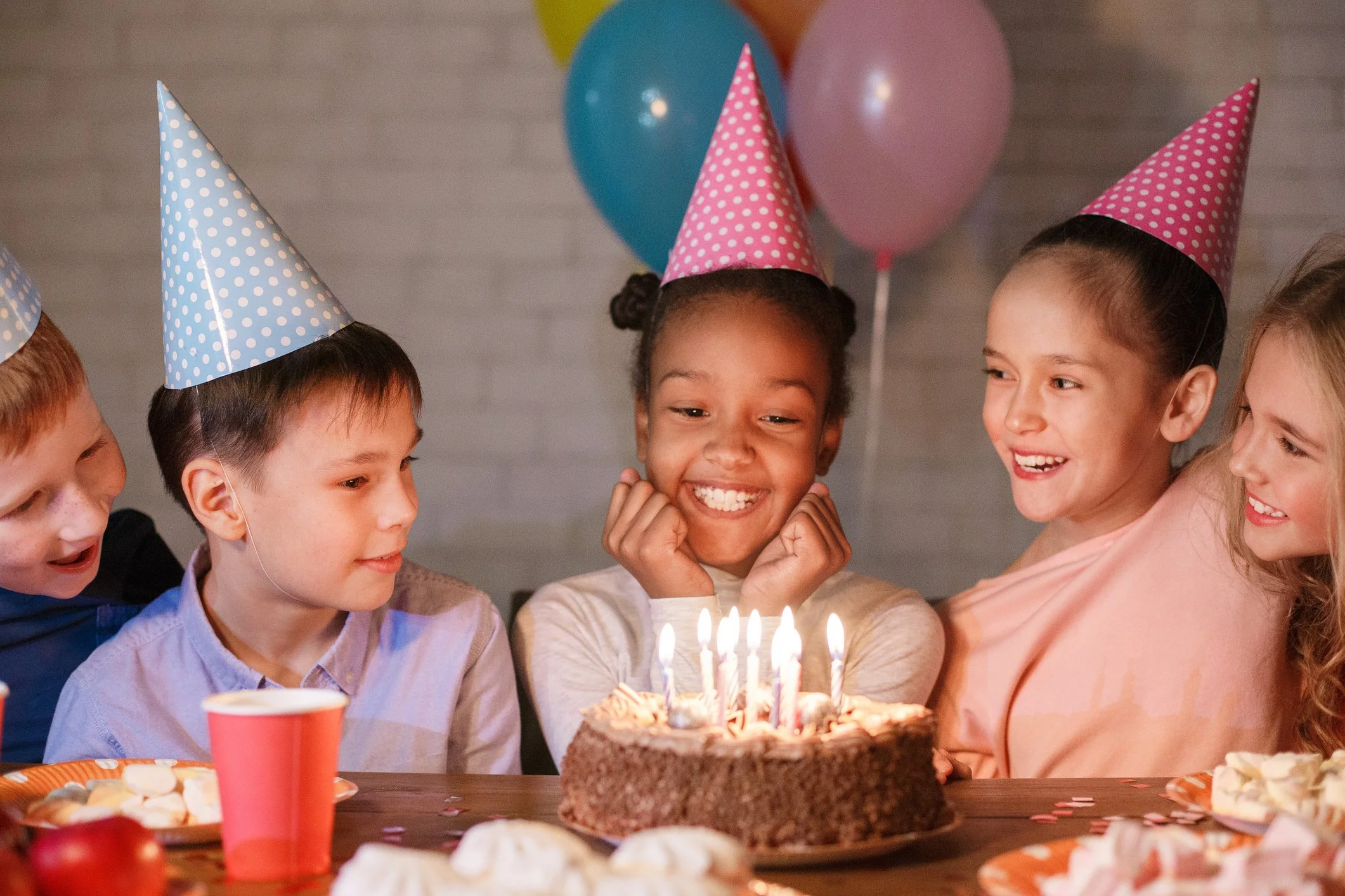 Group of children celebrating a birthday party with colorful balloons, wearing party hats, gathered around a chocolate cake with lit candles, smiling and laughing.