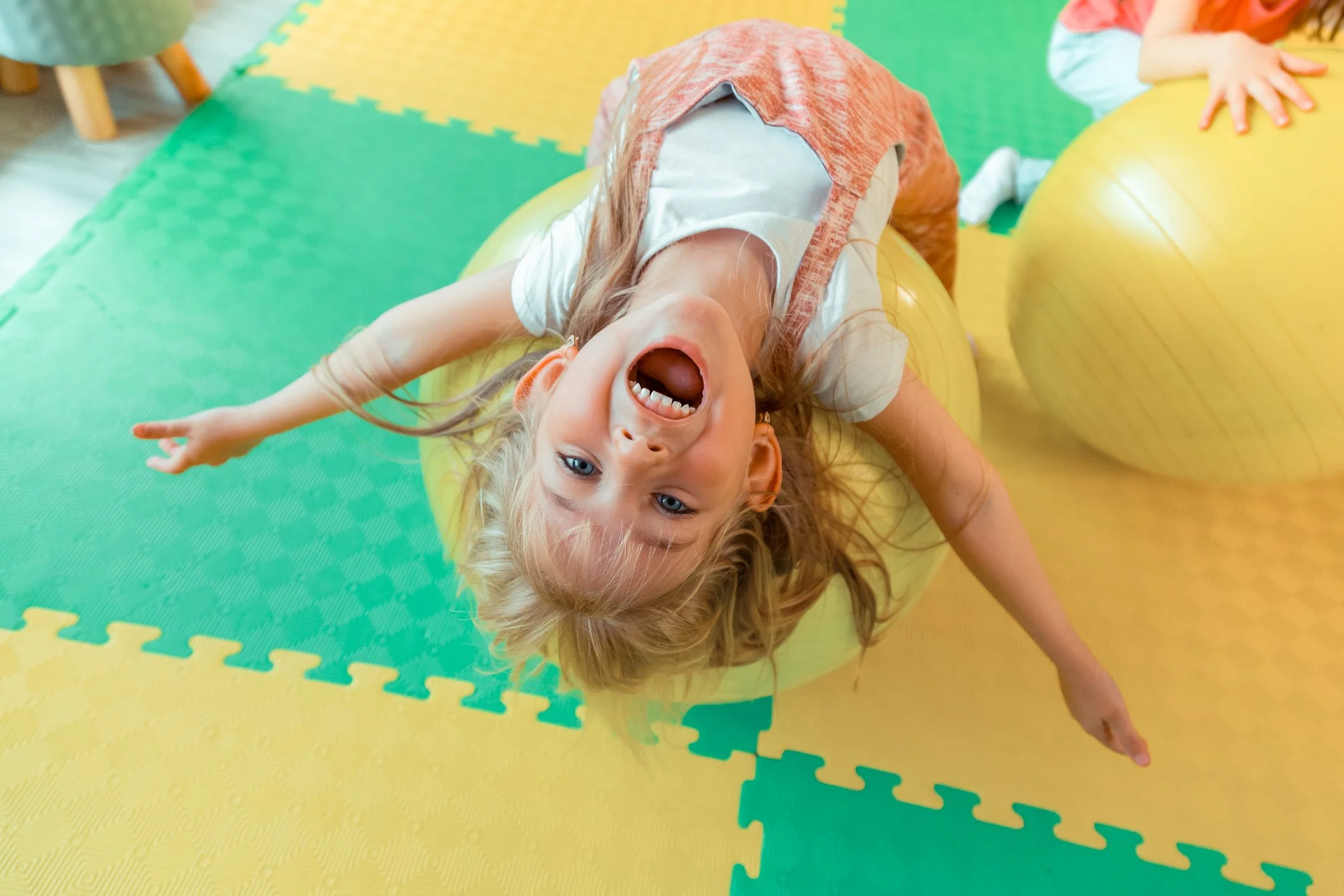 Child lying upside down on a large yellow exercise ball, with arms extended outward and hair hanging down, smiling with her mouth open, in a colorful indoor play area.
