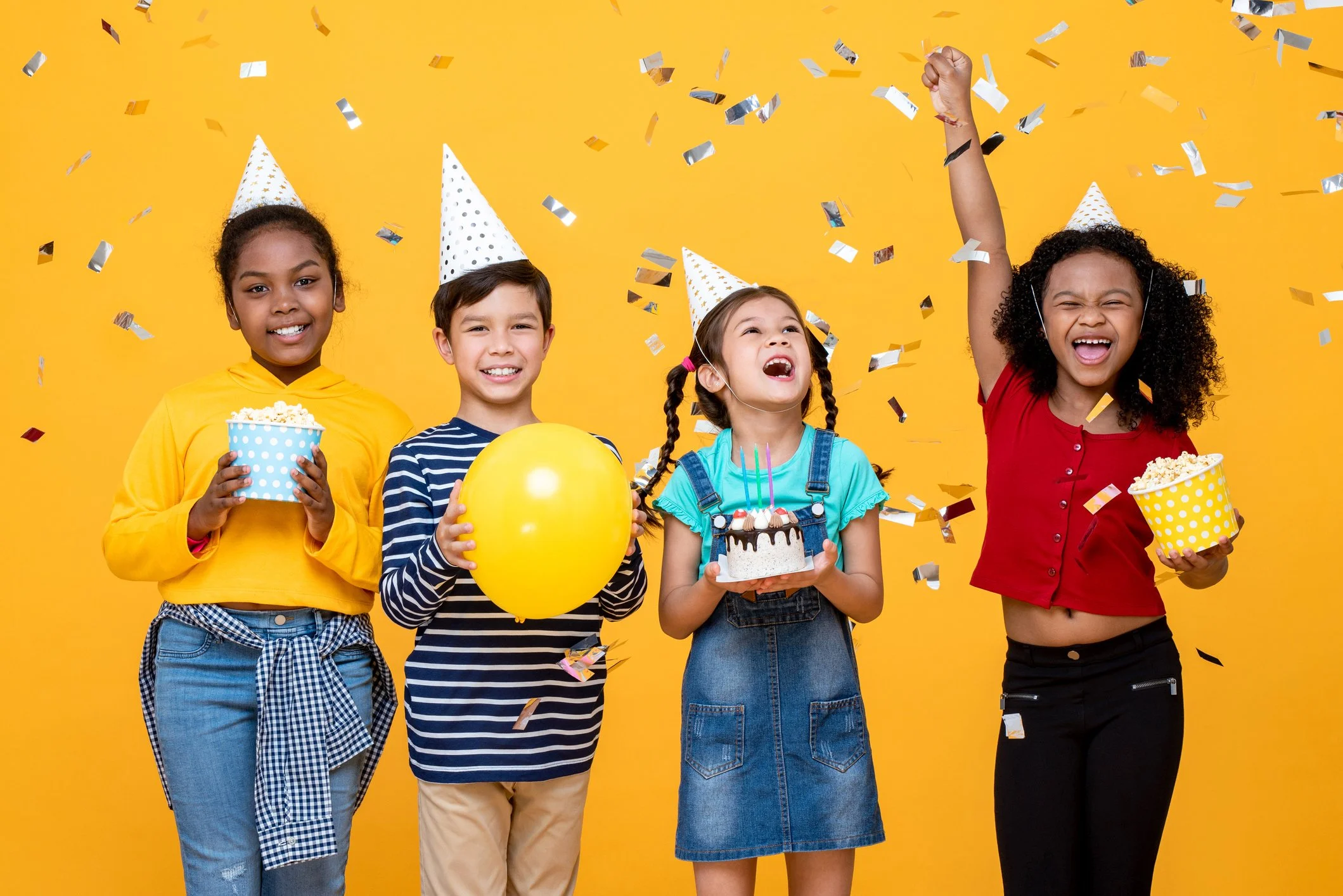 Children celebrating birthday party with balloons, cake, and confetti, wearing party hats, against a yellow background.
