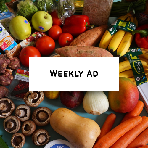 Fresh vegetables and fruits including apples, tomatoes, bell peppers, sweet potatoes, carrots, mushrooms, garlic, and a peach on a table with a sign that says 'Weekly Ad'.