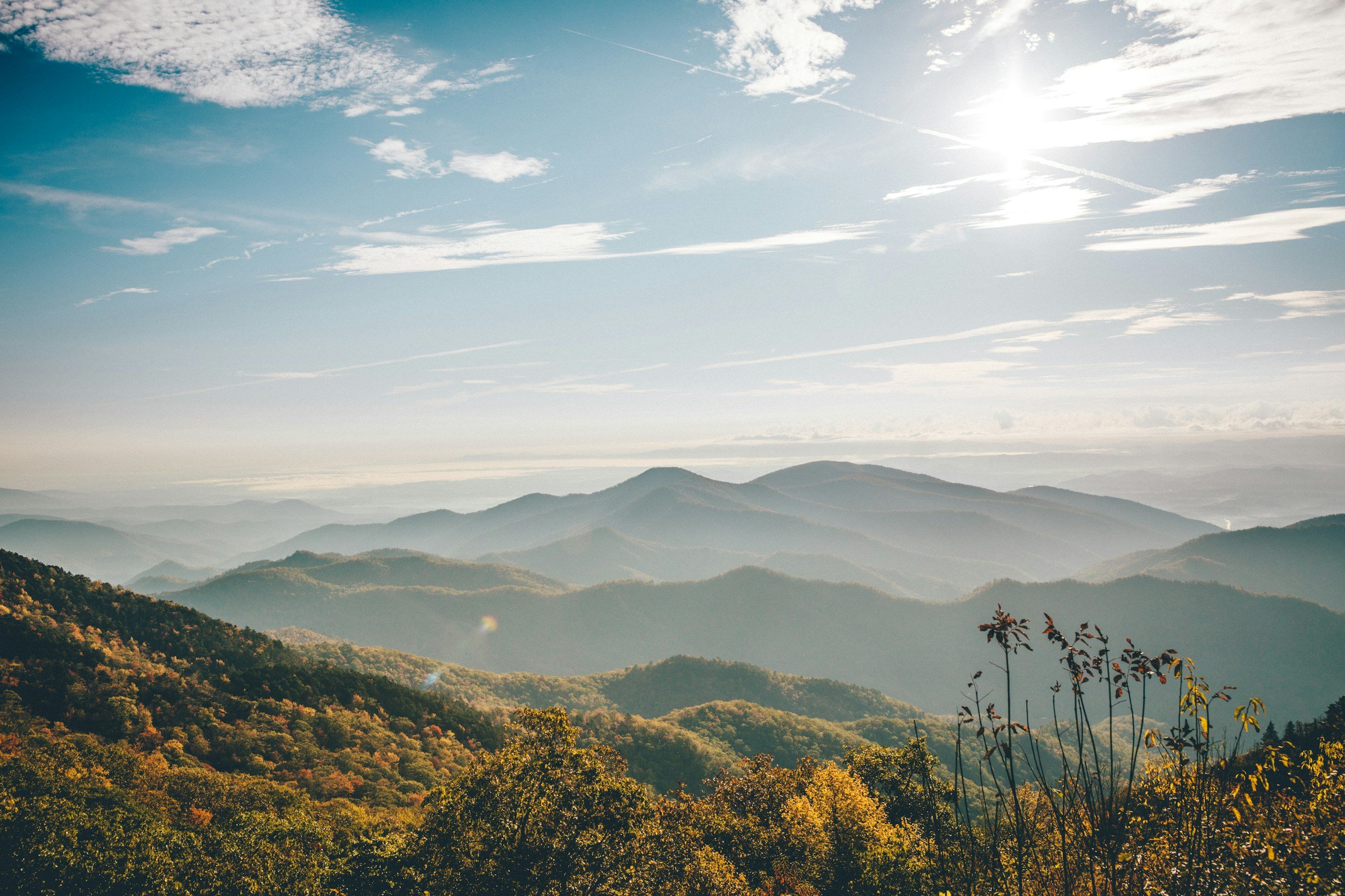 Landscape of mountain ranges with trees in autumn colors and a partly cloudy sky with the sun shining.