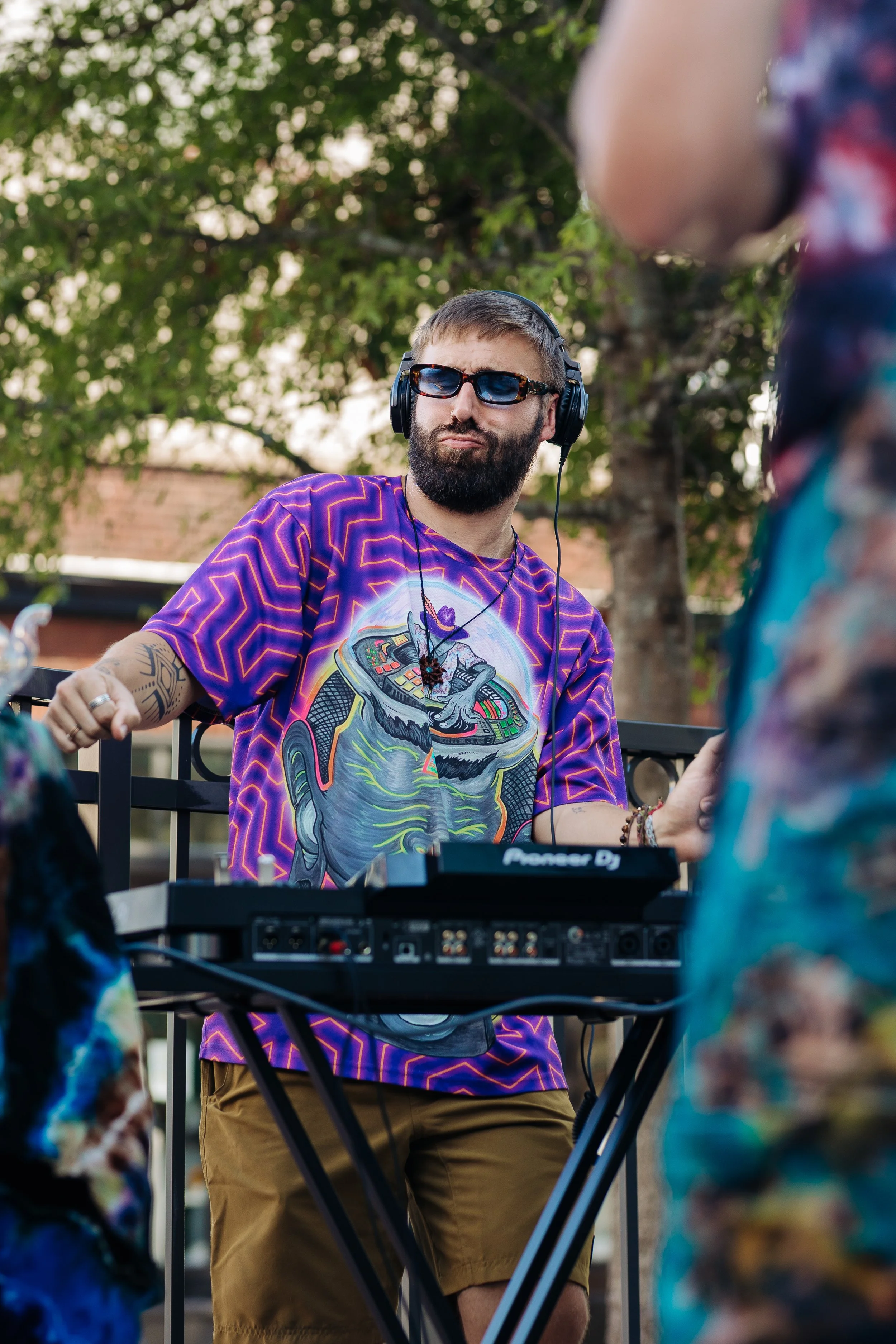 A DJ with a beard, wearing sunglasses and a colorful graphic t-shirt, is performing at an outdoor event. He is using a Pioneer DJ mixer and wearing headphones, with a tree and brick building in the background.