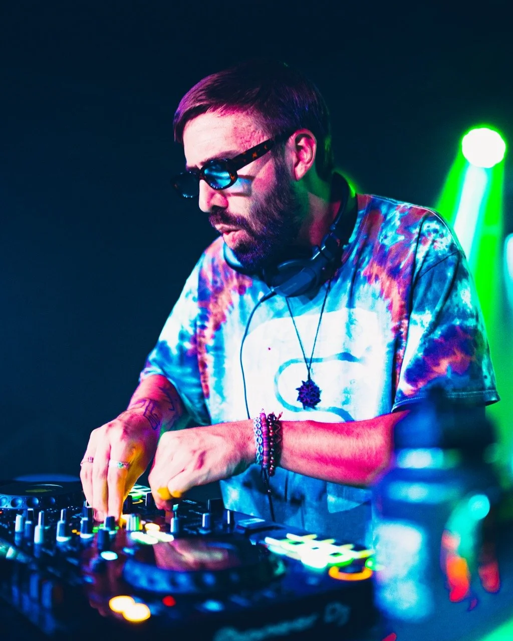 A DJ with glasses and a beard, wearing a tie-dye shirt, mixing music on a DJ console in a club with colorful lights.