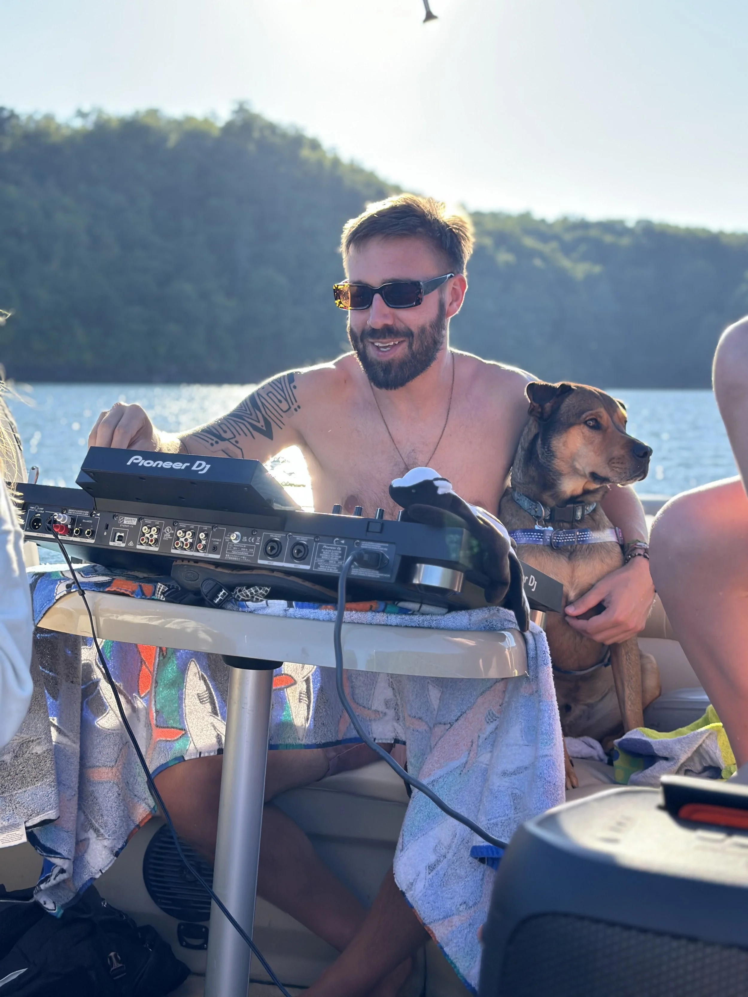 A man with sunglasses and a beard, shirtless, sitting at a table on a boat, with a dog on his lap, near a body of water with trees in the background, while DJ equipment is on the table.
