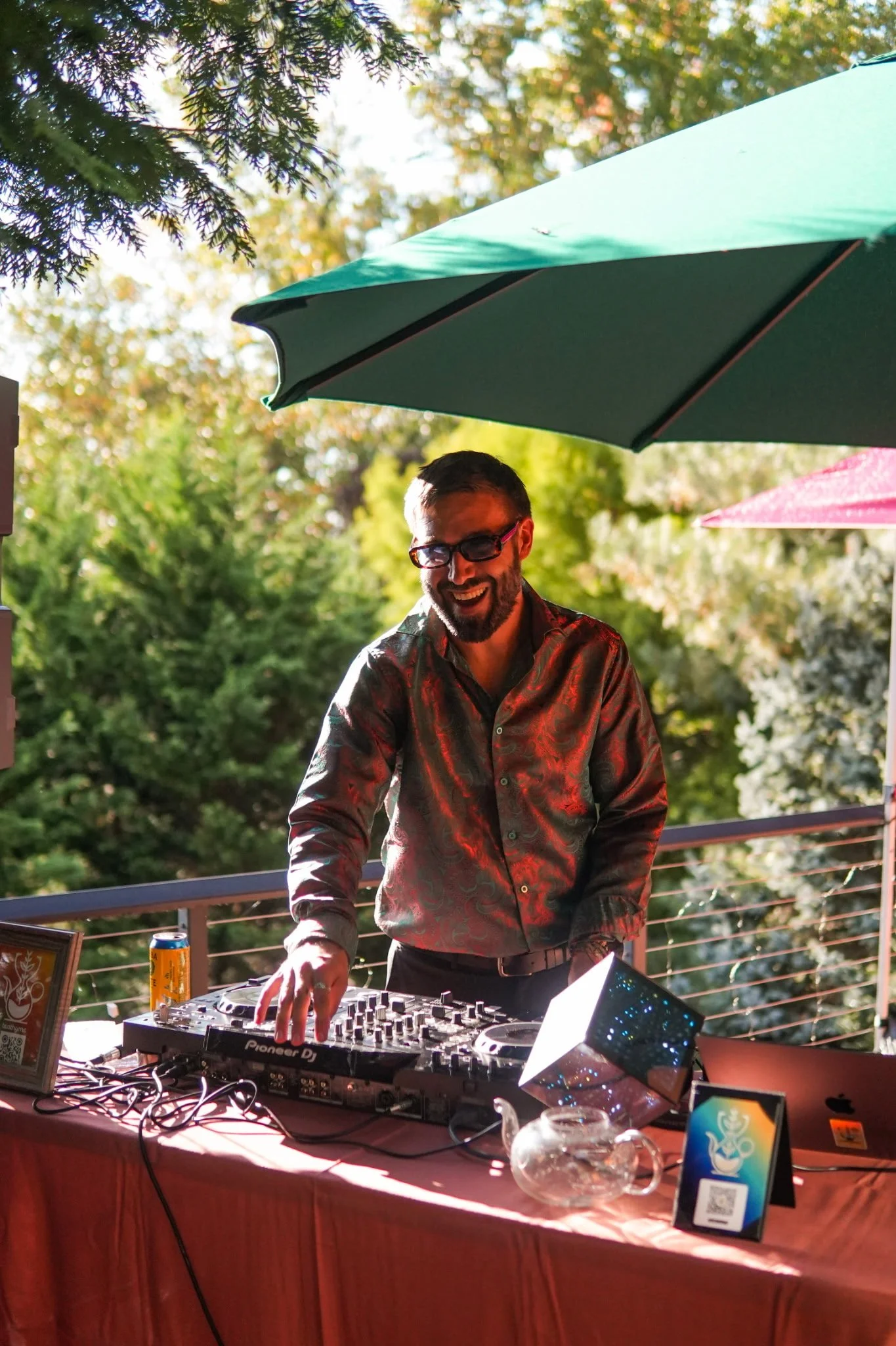 A man with glasses and a patterned shirt is DJing outdoors at a party or event with greenery in the background, under large umbrellas, smiling while operating DJ equipment on a table covered with a red tablecloth.