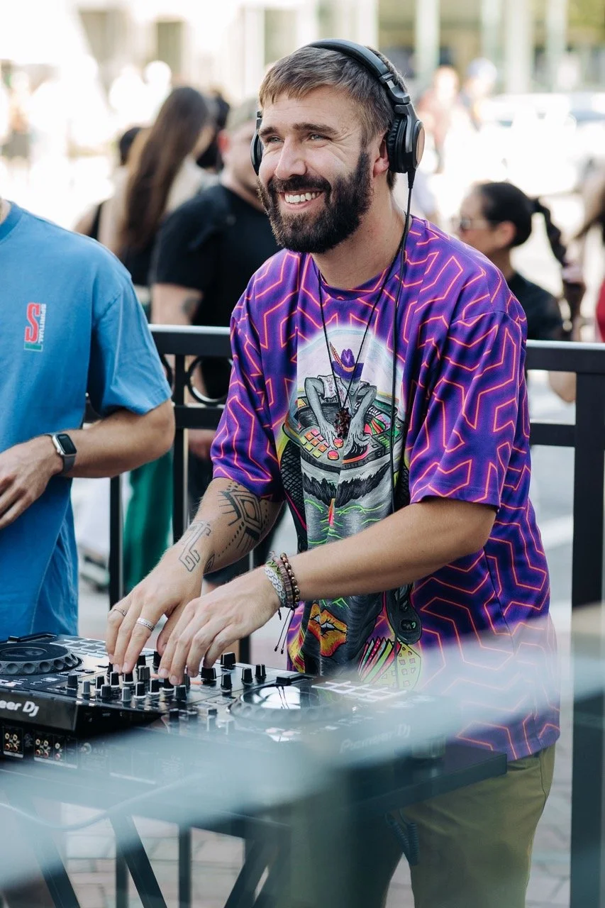 A man with a beard wearing headphones and a colorful graphic t-shirt is DJing outdoors surrounded by people.
