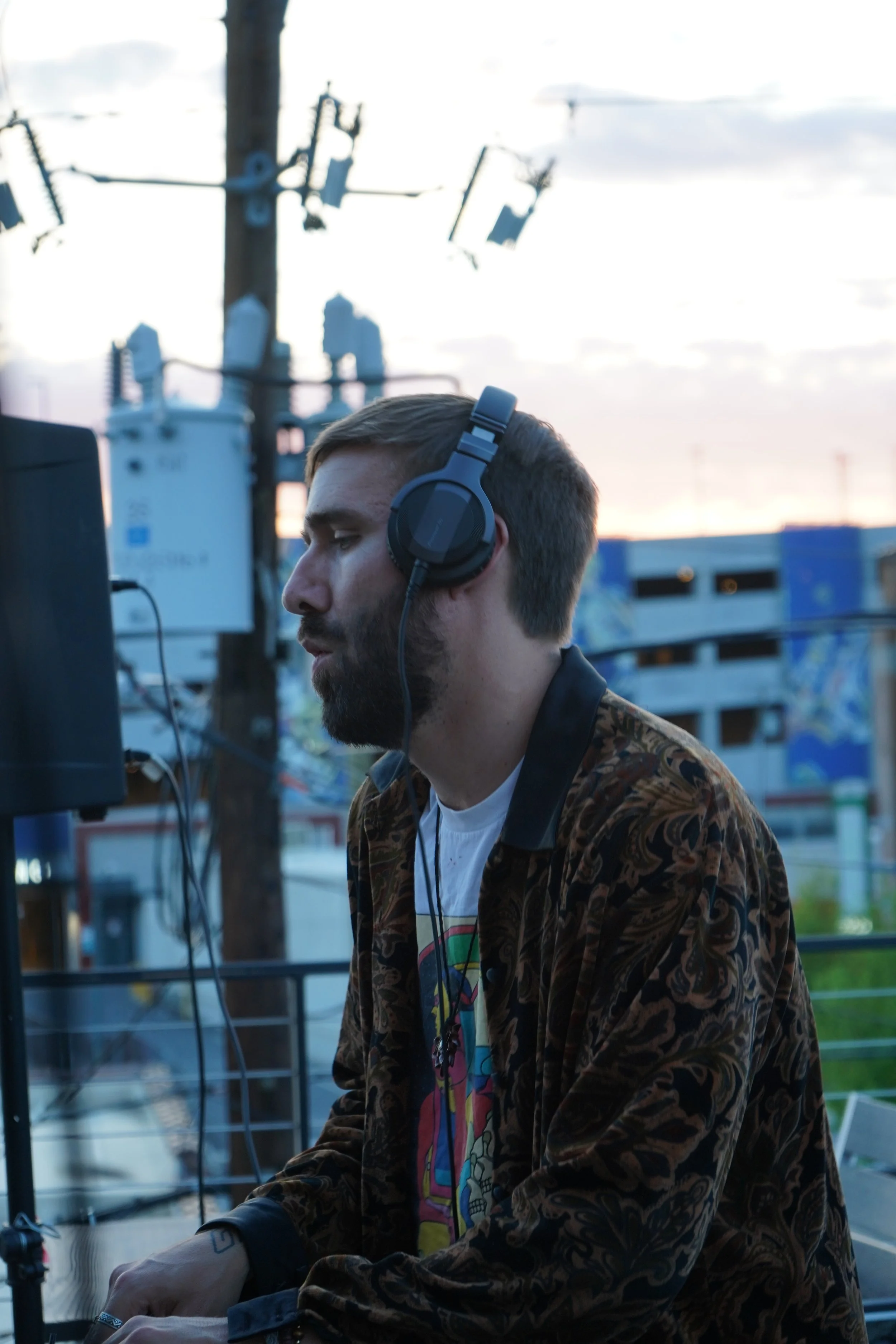 A man with a beard wearing a patterned shirt and headphones DJing outdoors during sunset.