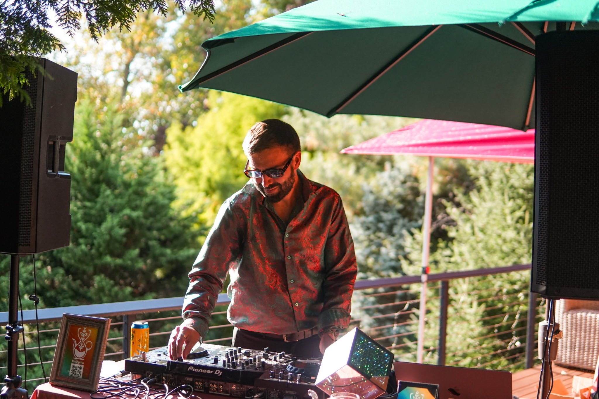 A man with sunglasses DJing at an outdoor event, with a table of equipment, framed picture, and colorful umbrellas in a garden setting.