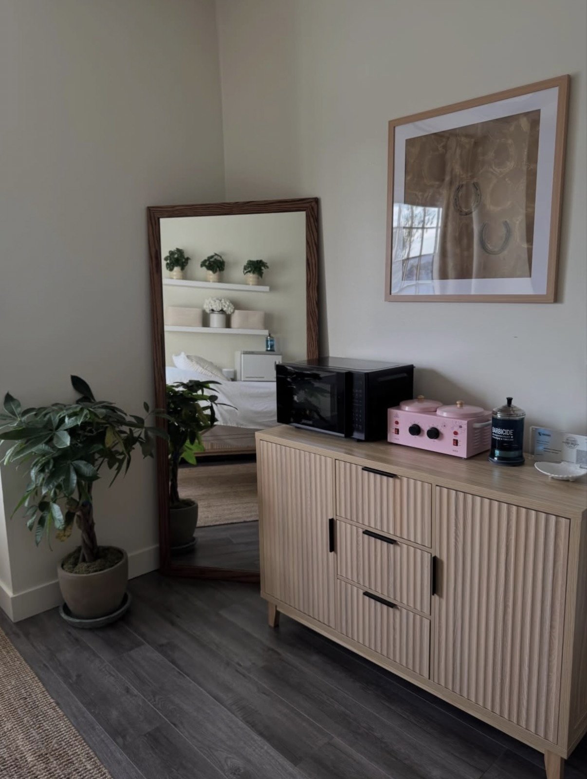 Interior of a bedroom with a wooden dresser on top of which is a microwave, a pink kitchen appliance, and cleaning supplies. A large mirror leans against the wall, reflecting a bed with a white headboard and wall-mounted shelves with plants and decorative items. A framed artwork hangs on the wall above.
