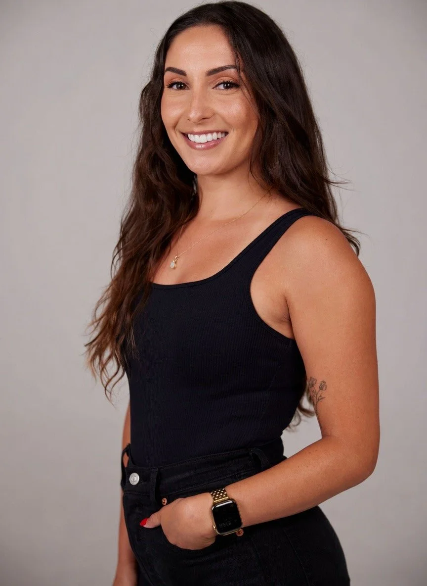 A woman with long brown wavy hair smiling and wearing a black sleeveless top, black jeans, a gold watch, and a delicate necklace, standing against a neutral background.