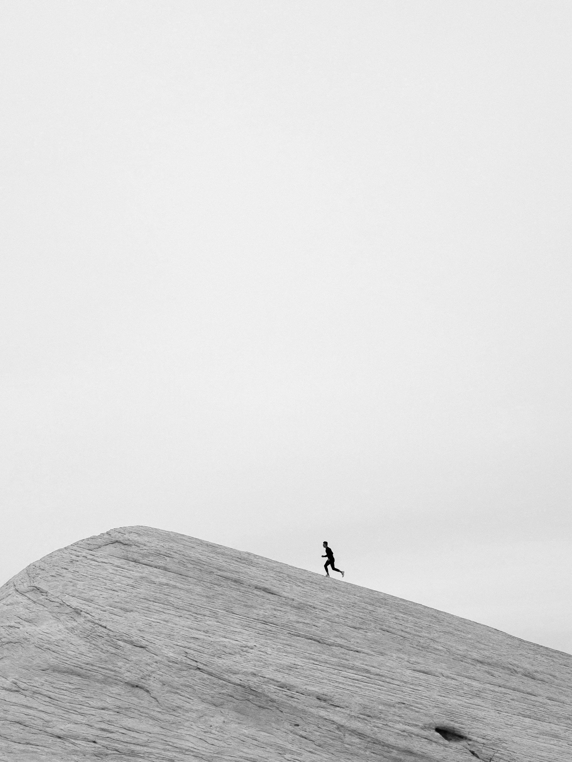 A person running on a large, smooth, sloped rock formation with a plain, light gray sky in the background.