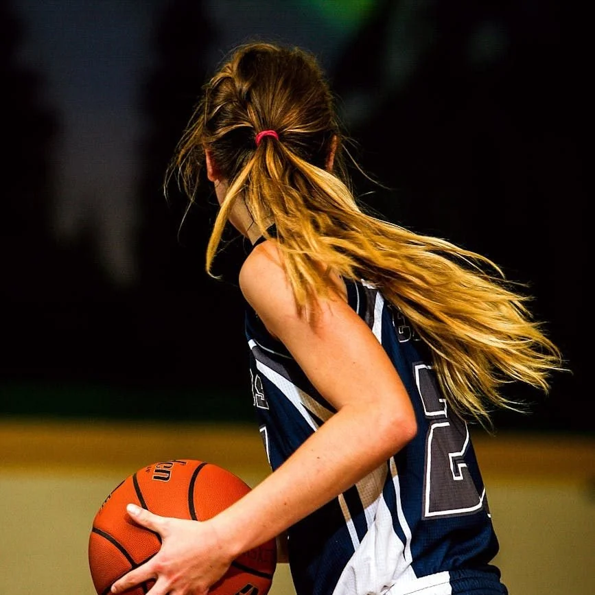A young girl with long blond hair in a ponytail, wearing a basketball uniform, holding a basketball on a court.
