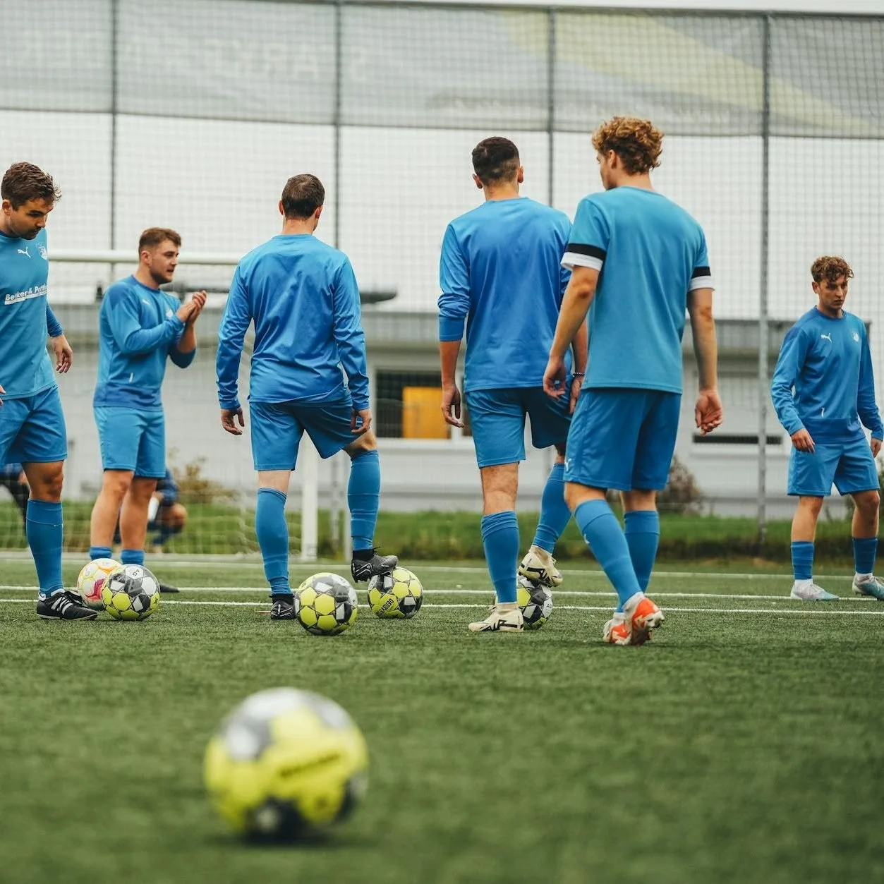 Group of soccer players in blue uniforms on a field with several soccer balls at their feet.