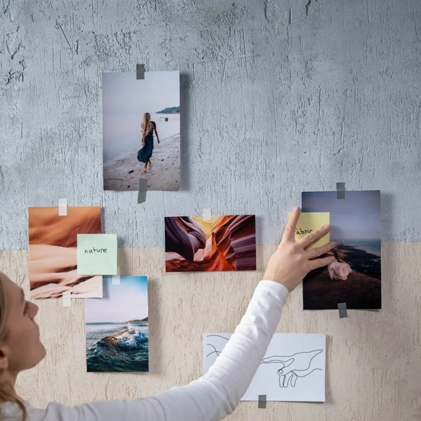 Person arranging photographs and notes on a textured wall, including images of a woman on a beach, ocean waves, canyon formations, and a landscape at sunset.
