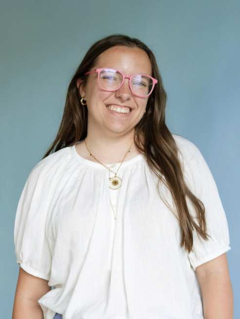 A woman with long brown hair, pink glasses, and gold jewelry, smiling against a blue background.