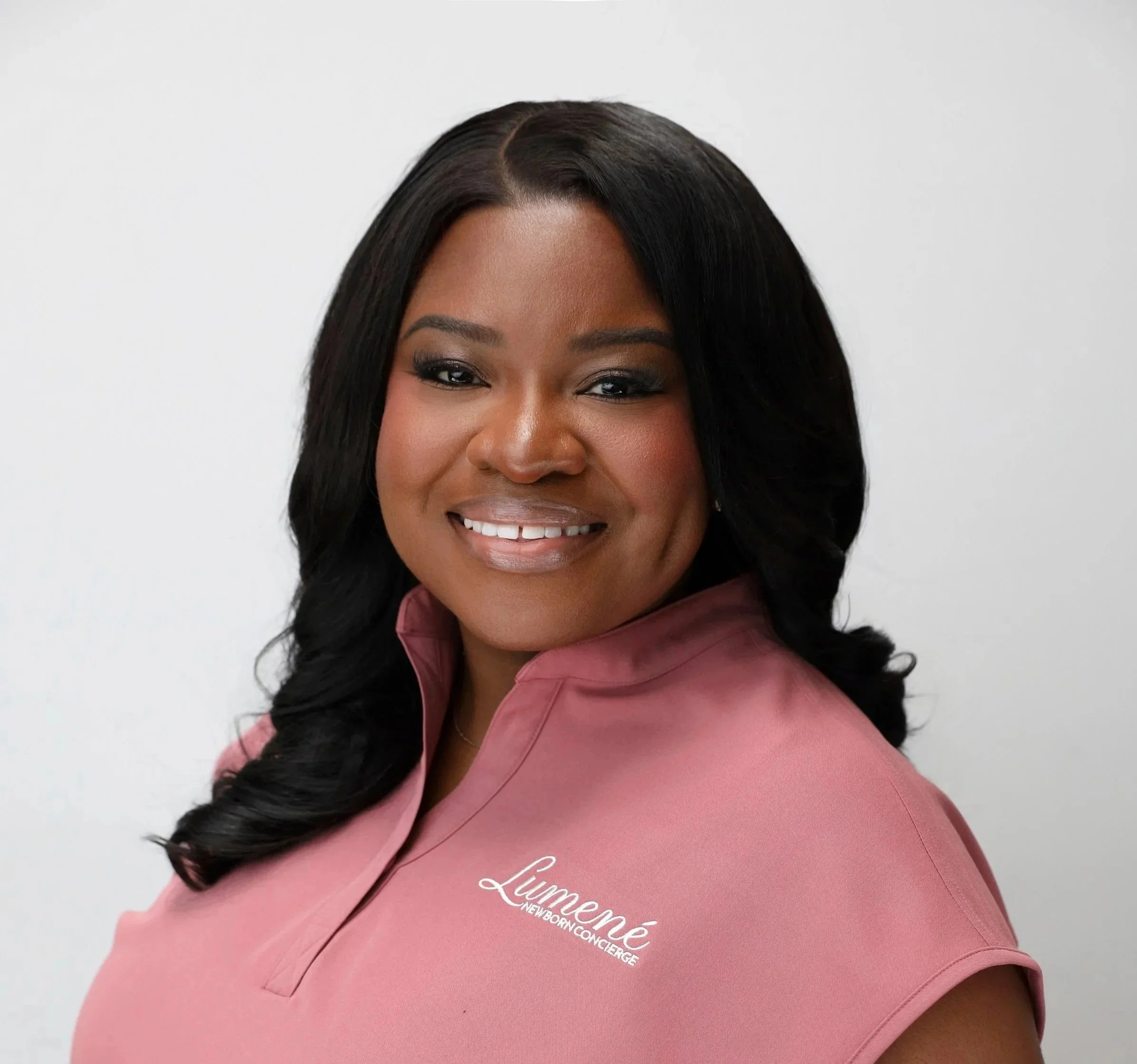 Close-up of a woman with long black hair and dark eye makeup smiling at the camera, wearing a pink uniform with white text, standing against a white background.
