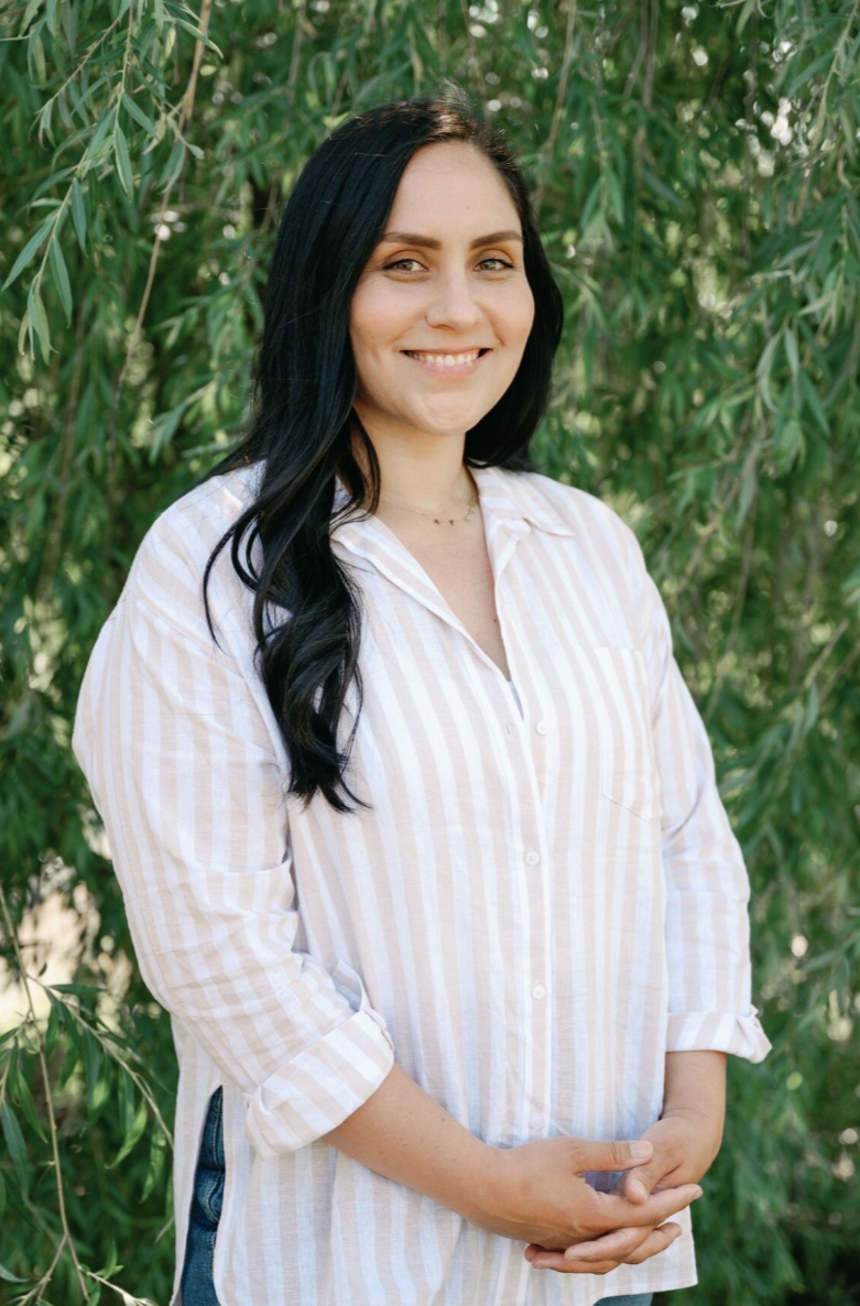 A woman with long black hair smiling outdoors in front of green leafy trees, wearing a light-colored striped button-up shirt.