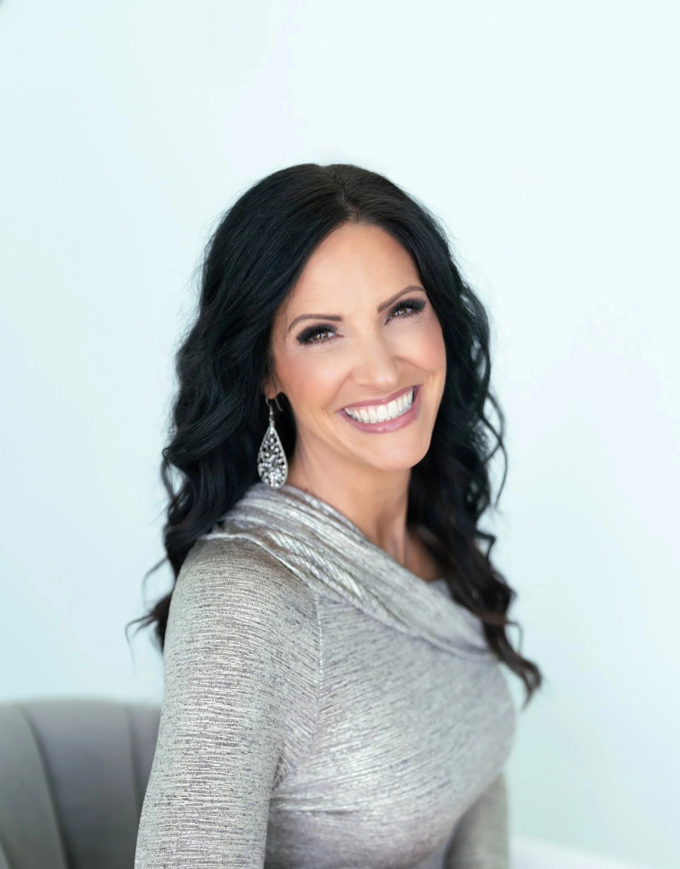 A woman with black hair, wearing makeup with eye shadow, mascara, and lipstick, smiling and wearing a silver, shimmery dress with large earrings, against a plain light background.