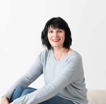 Woman with black hair and a light gray sweater, sitting on a beige surface, smiling against a plain white background.
