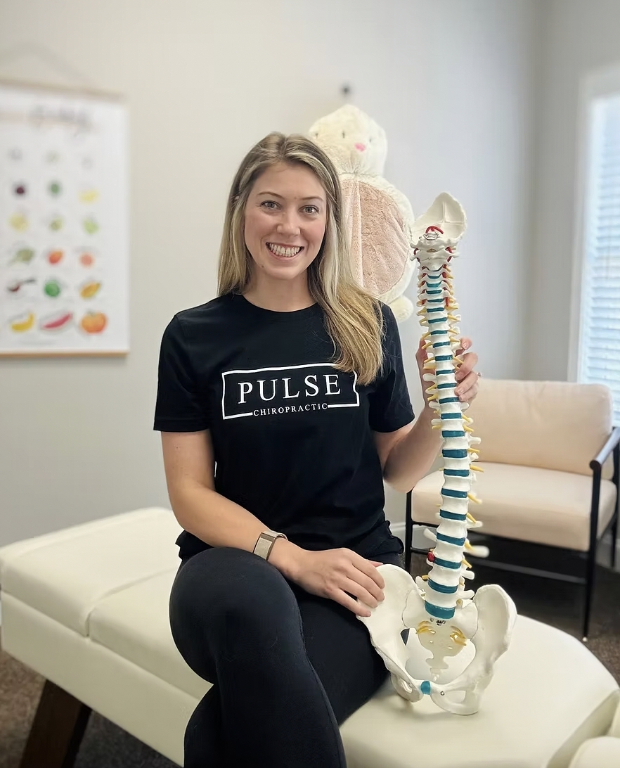 Woman sitting on a medical examination table holding a model of the human spine, wearing a black Pulse Chiropractic T-shirt, with a plush butterfly on the wall behind her.