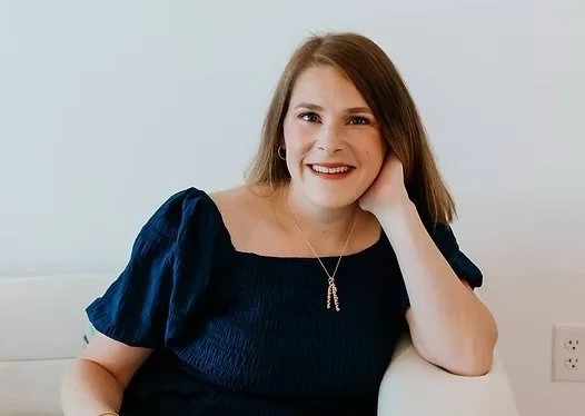 A smiling woman with shoulder-length brown hair wearing a dark blue top and a necklace, sitting against a light background.