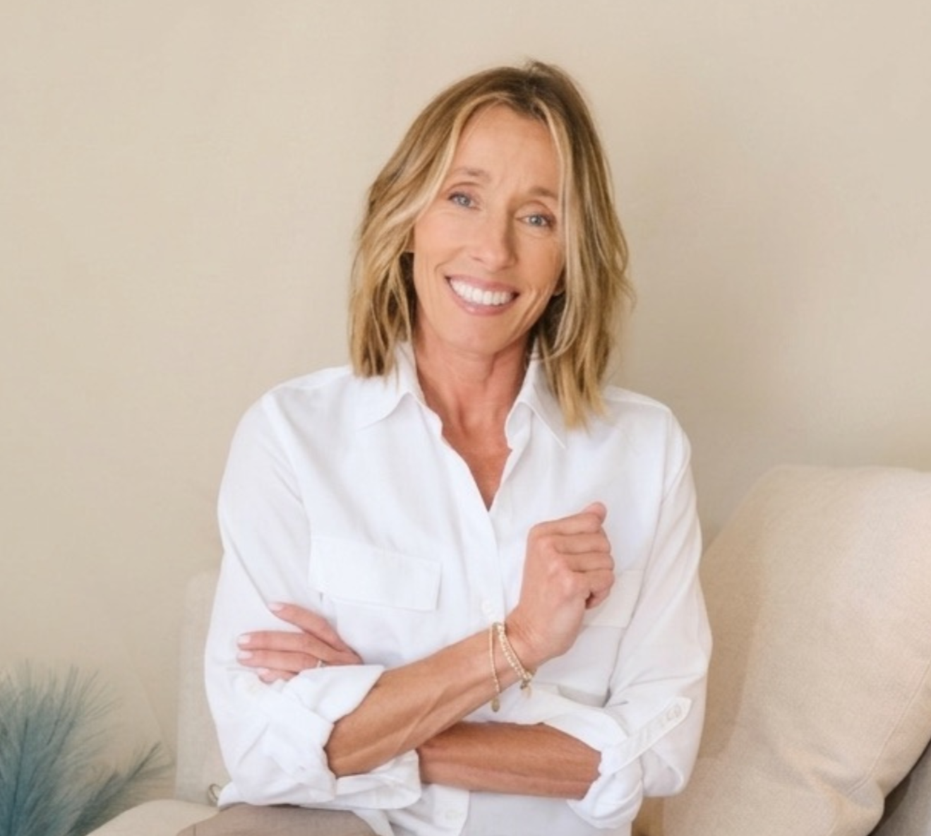 A smiling woman with shoulder-length blonde hair wearing a white button-down shirt, standing with arms crossed in a living room with beige walls and furniture.