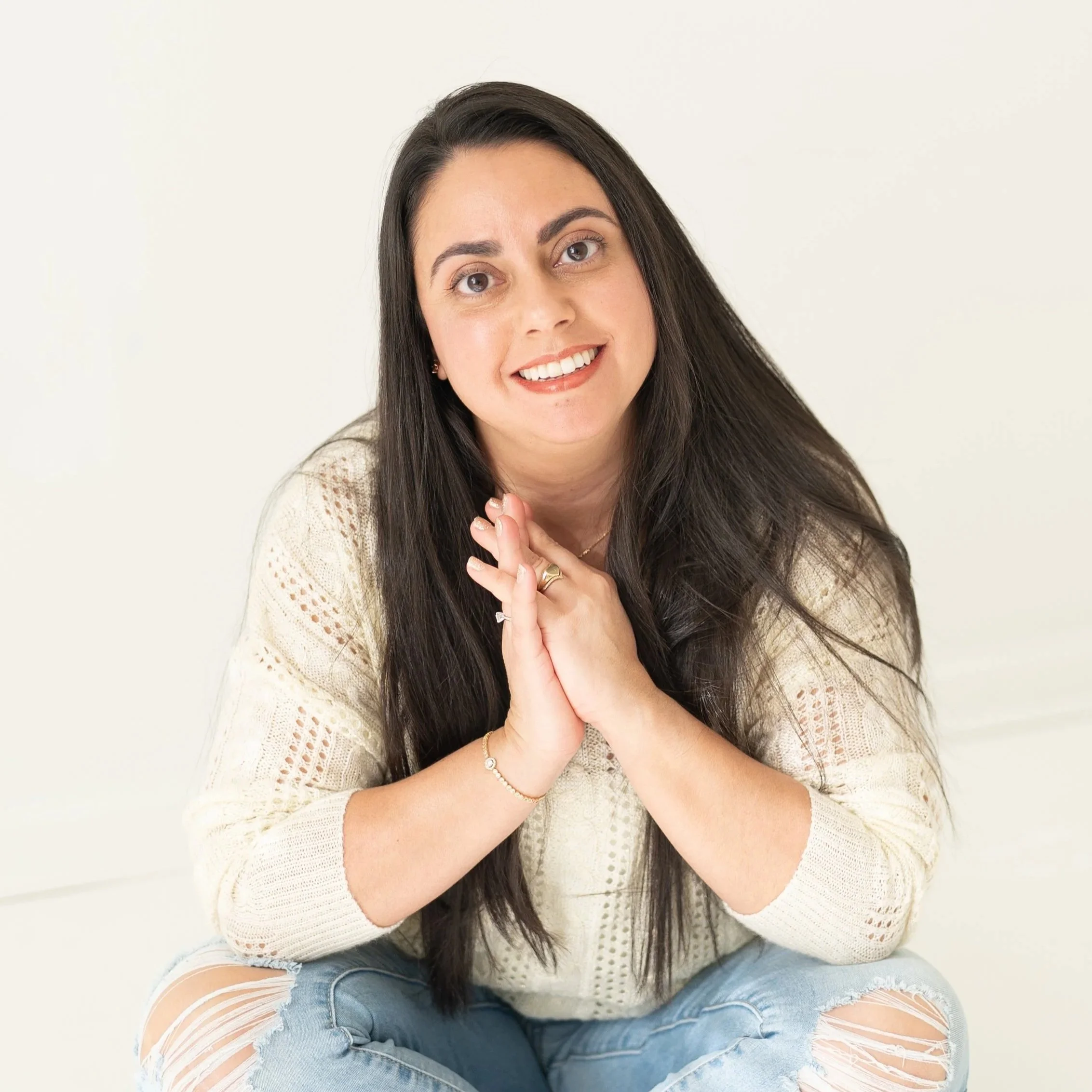 A young woman with long dark hair, smiling and looking at the camera, sitting against a plain light-colored background. She is wearing a cream-colored knitted sweater and ripped blue jeans.