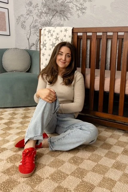 Young woman sitting on a patterned carpet next to a wooden crib, smiling at the camera, with a mint green sofa and decorative pillows in the background.