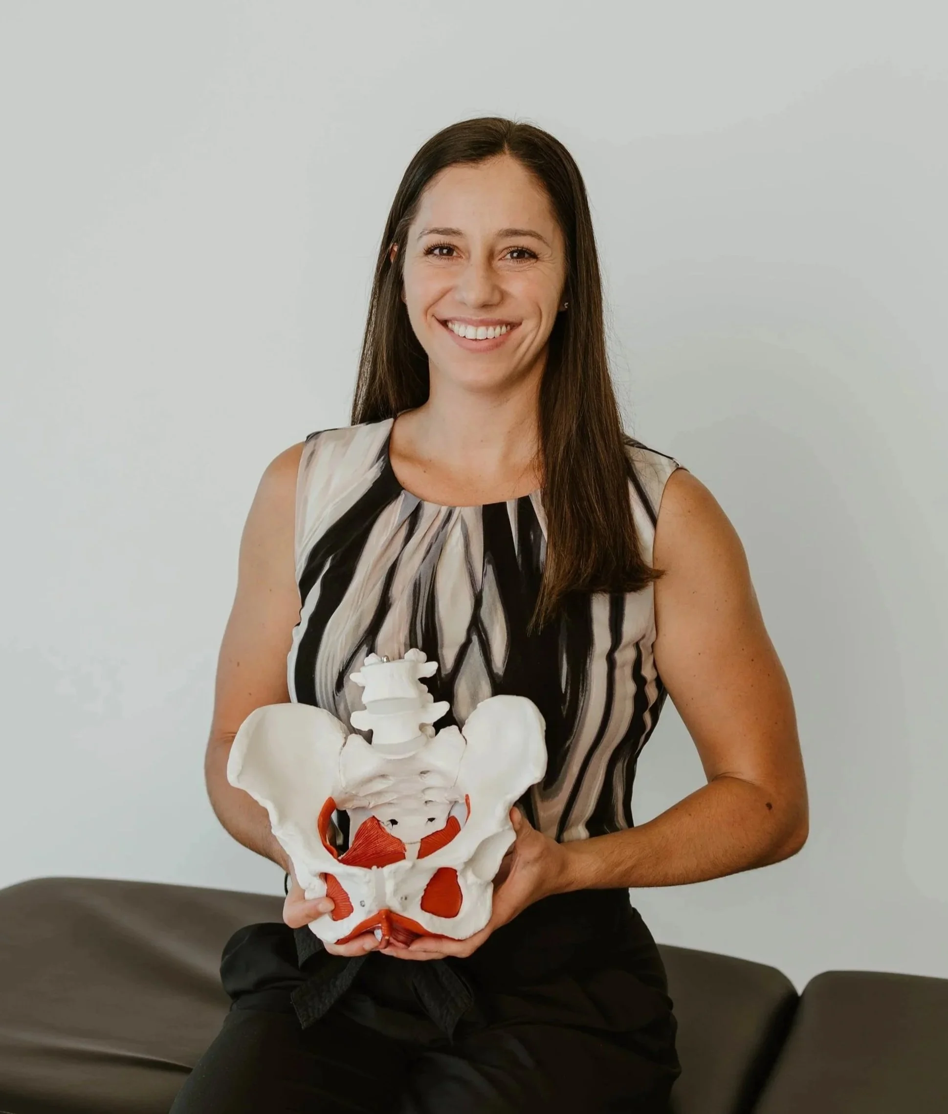 Woman smiling and holding a large anatomical model of a pelvis in front of her chest, sitting on a bench against a plain light-colored wall.