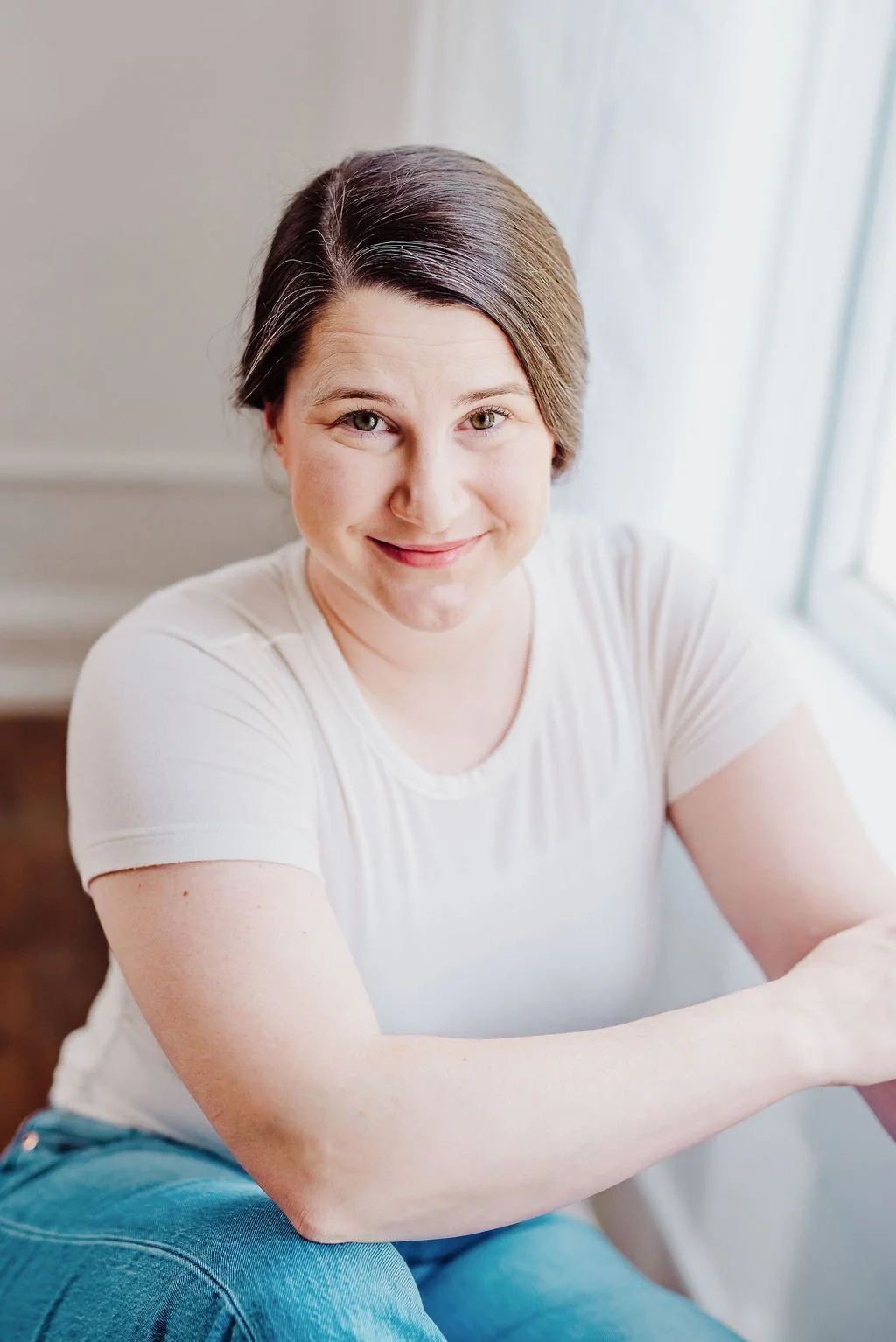 A woman with short brown hair, wearing a white t-shirt, smiling at the camera, sitting near a window with light streaming in.