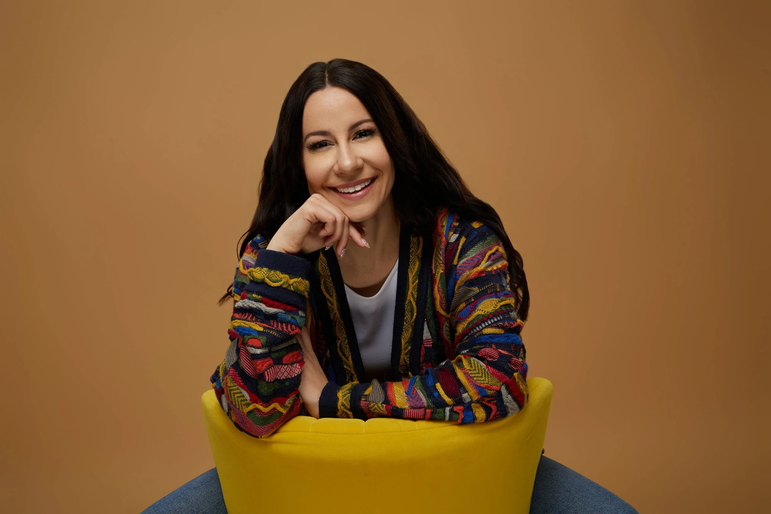 A woman with long dark hair smiling, sitting in a yellow chair with one hand resting on her chin, wearing a colorful patterned sweater against a tan background.
