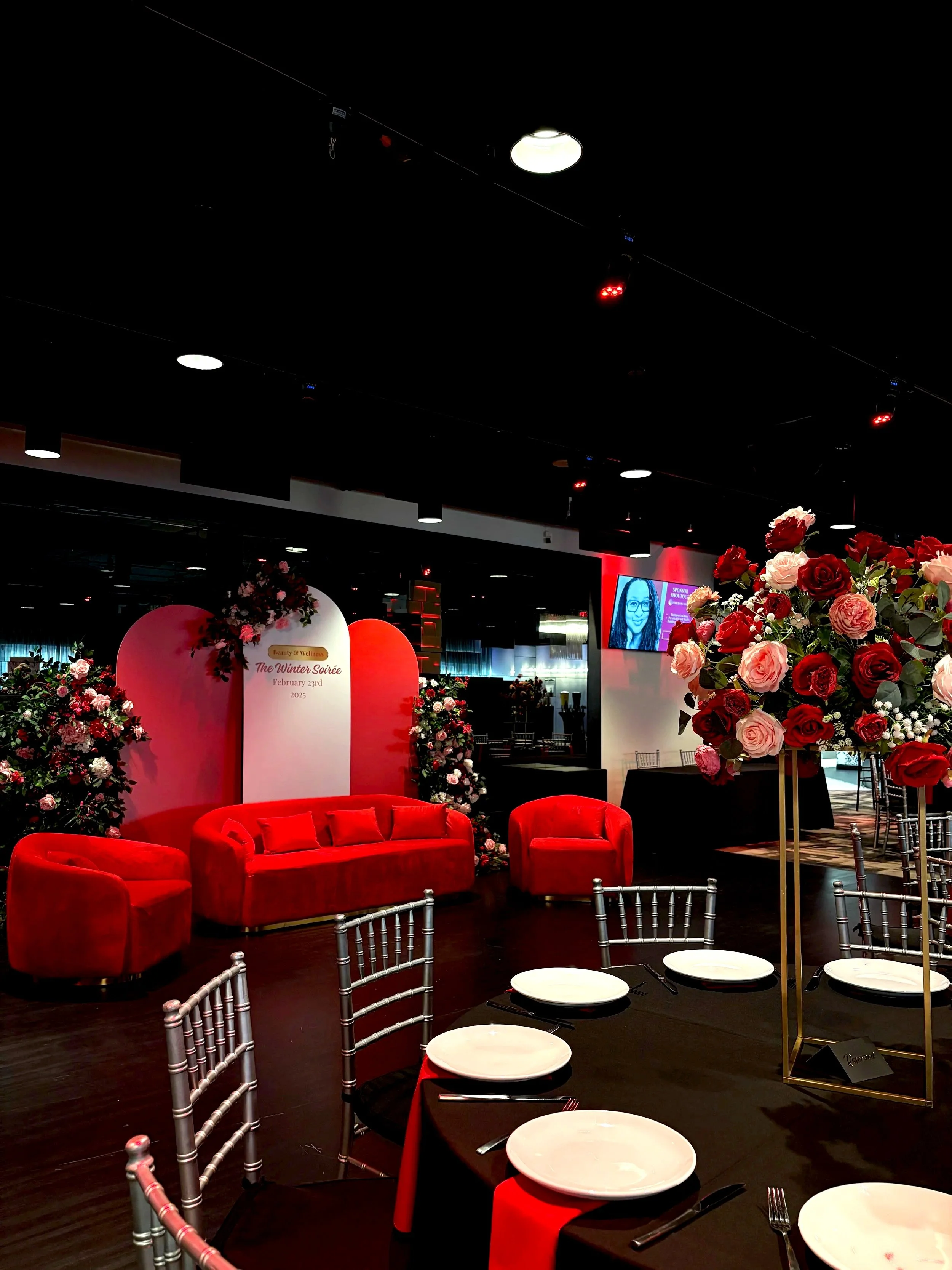 Venue decorated for a celebration with a pink and red theme, featuring a red velvet sofa, chairs, and floral arrangements of pink and red roses, set around tables with white plates and black napkins, with a monitor screen displaying an image of a woman.