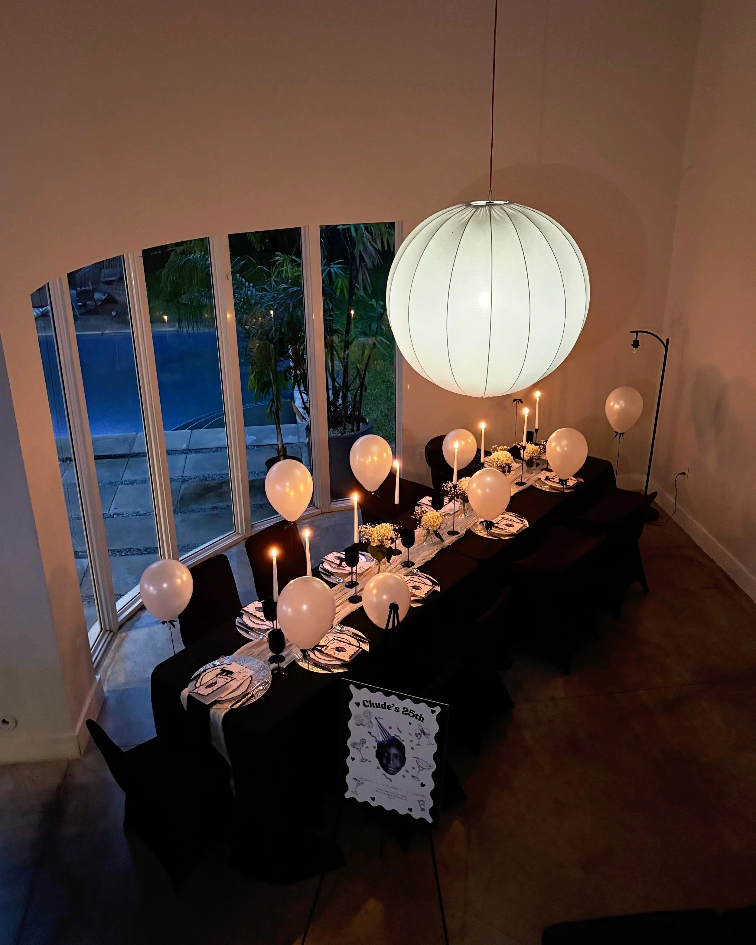 A long dining table decorated with white balloons, lit candles, flowers, and party plates, set inside a room with large windows overlooking a backyard pool.