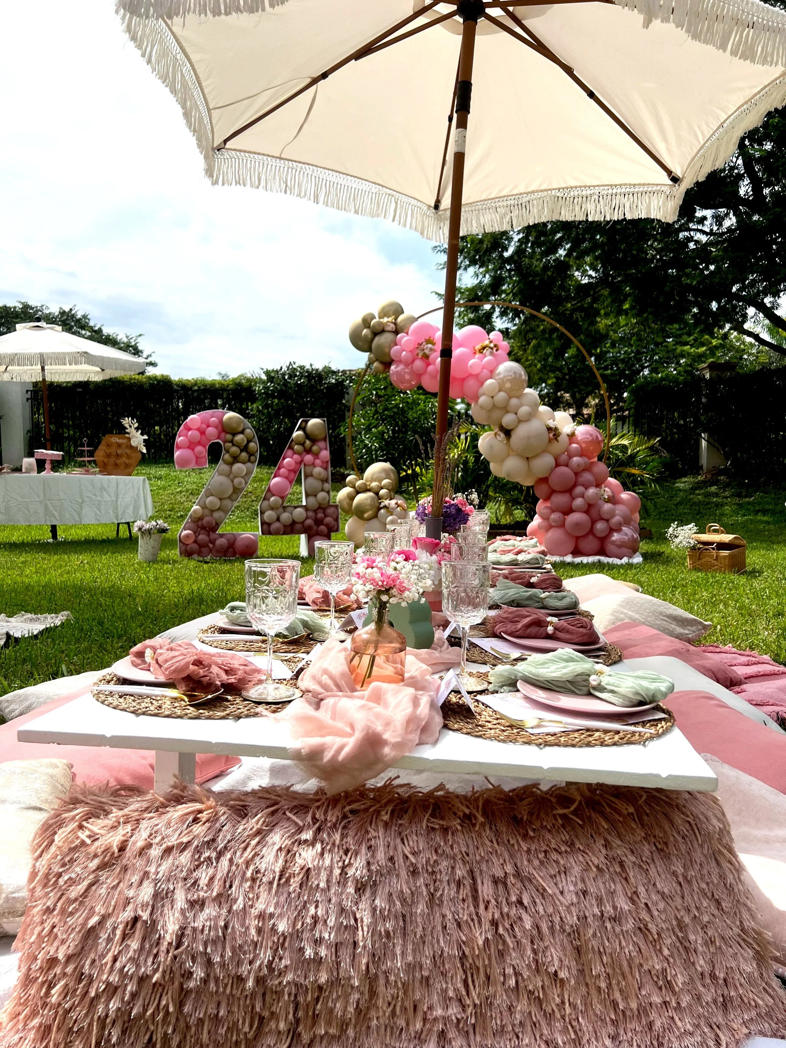 Decorated outdoor table for a celebration, with pink, white, and gold balloons, floral centerpieces, and set with glassware and napkins, under a large umbrella.