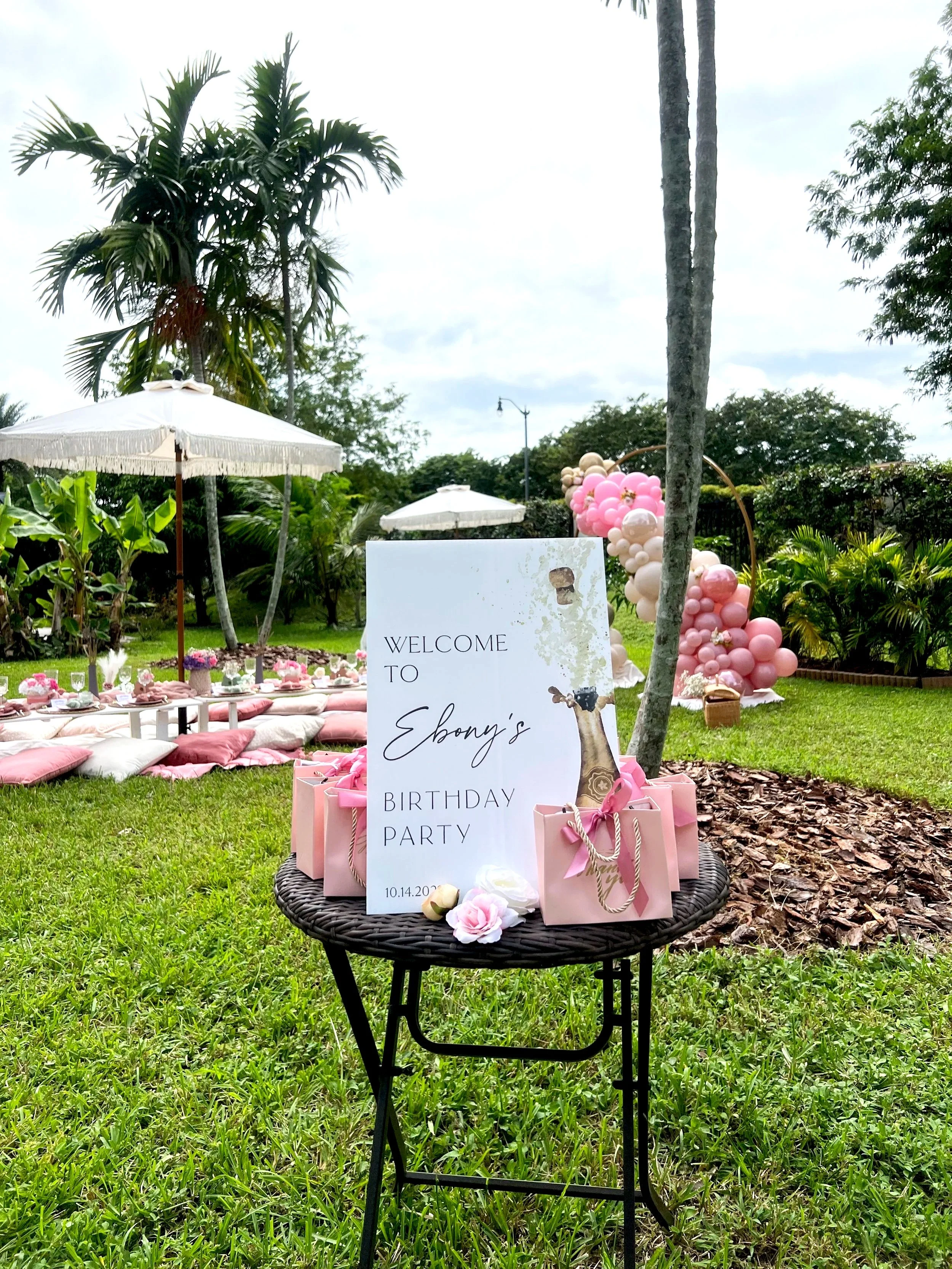 A newlywed couple sharing a tender moment outdoors, the groom gently leaning his forehead against the bride's, both smiling softly. The bride is wearing a lace wedding dress with short sleeves and a floral crown of greenery and small white flowers. The groom is dressed in a black suit with a white shirt and black tie, standing behind and slightly to the side of the bride in a natural, green setting.